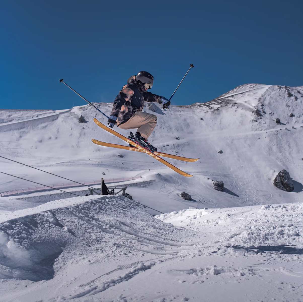 Uno sciatore salta a mezz'aria da una rampa di neve su un pendio di montagna sotto un cielo azzurro e limpido.