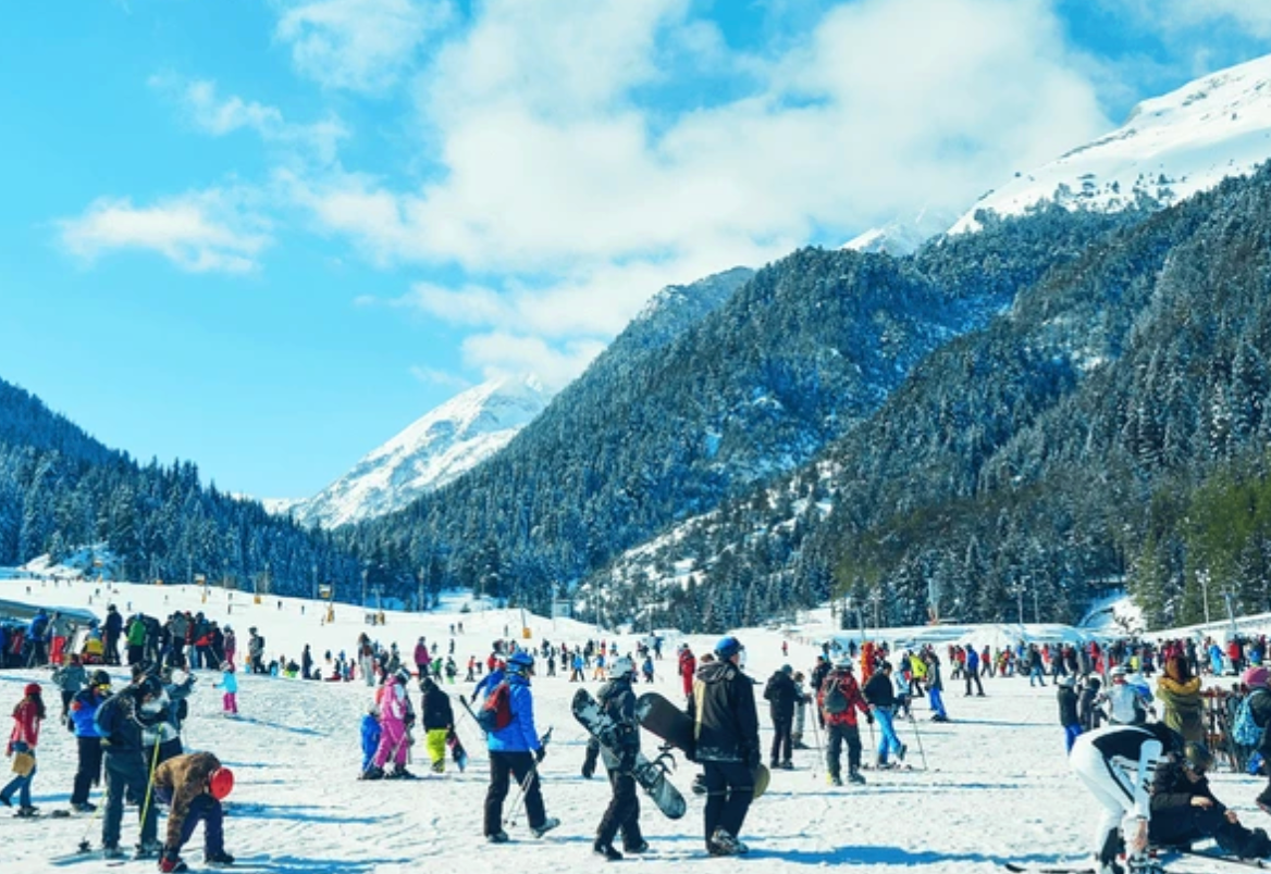 Numerosi sciatori e snowboarder su una pista affollata ai piedi di montagne innevate sotto un cielo blu.