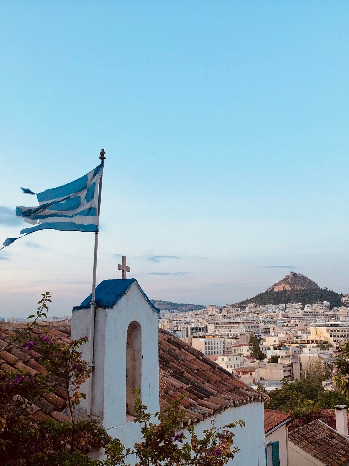 Una bandera griega ondea al viento sobre un campanario blanco con techo azul, con vistas a un denso paisaje urbano y una gran colina al anochecer.