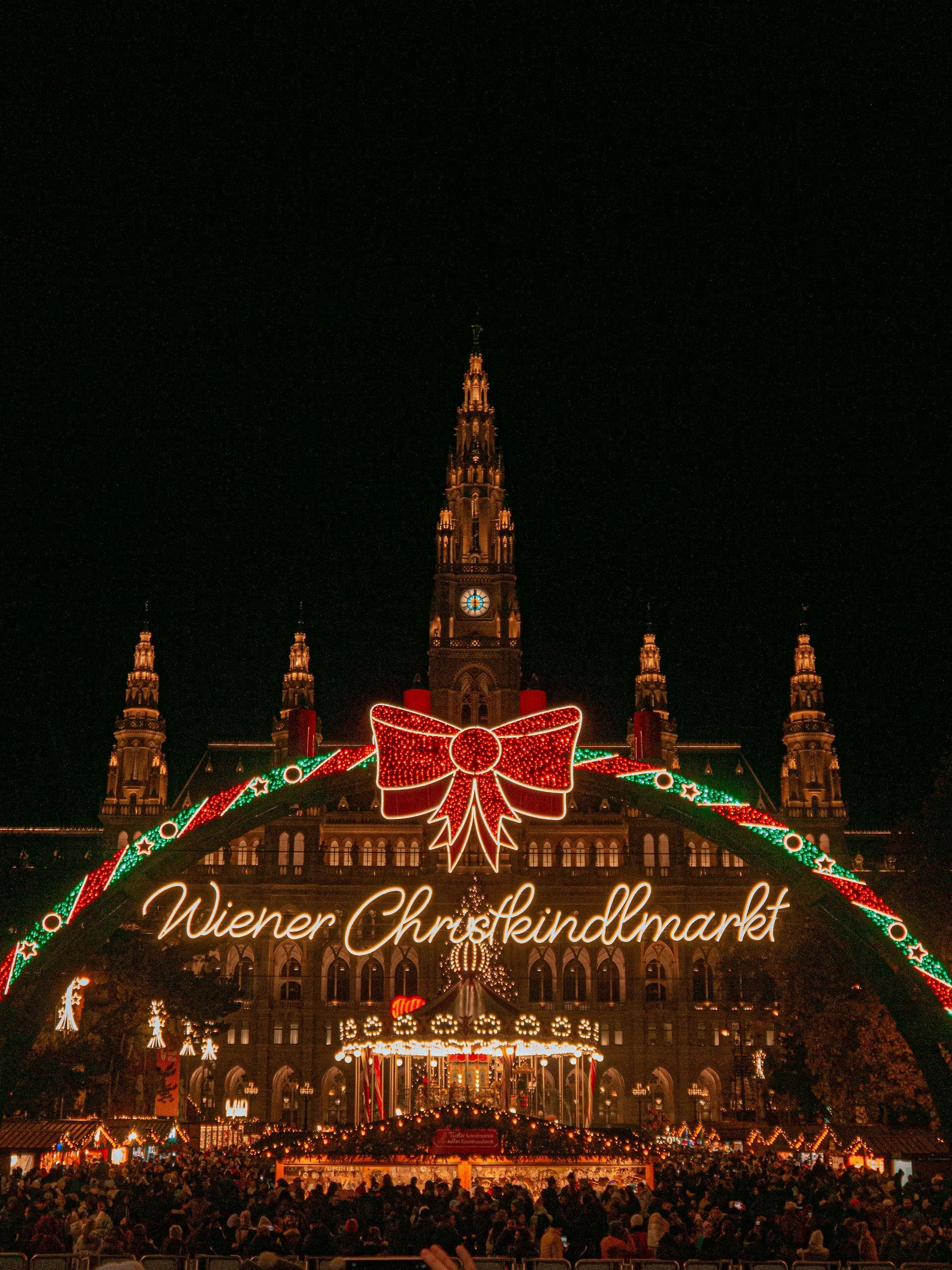 Der mit einer roten Schleife geschmückte Eingangsportalbogen des Wiener Christkindlmarktes erstrahlt nachts vor einem prunkvollen Gebäude, darunter eine Menschenmenge.