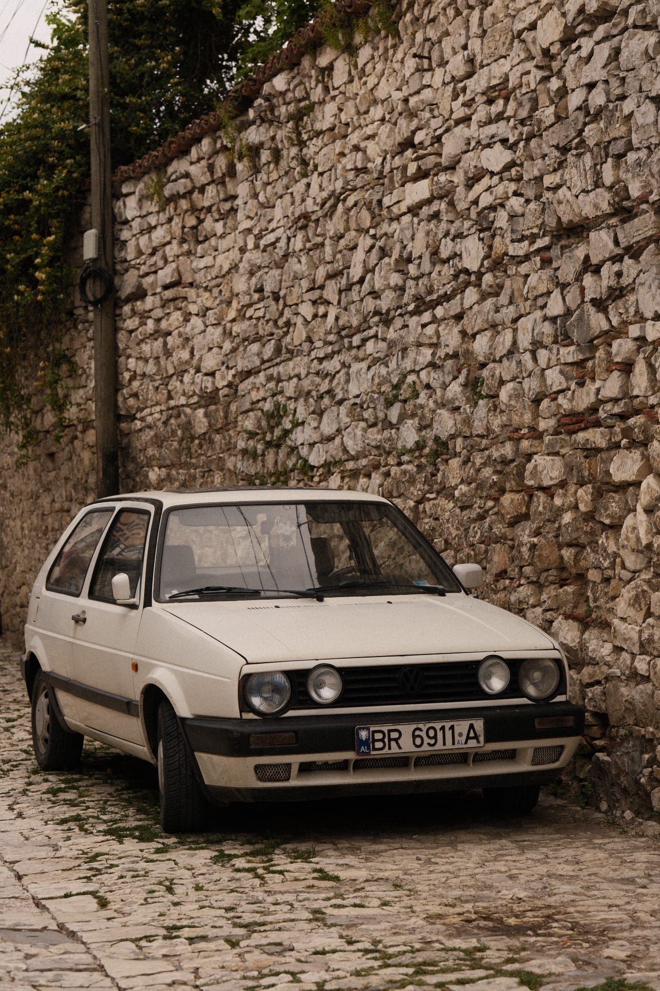 Un'auto bianca d'epoca parcheggiata su una strada acciottolata accanto a un alto muro di pietra rustico.