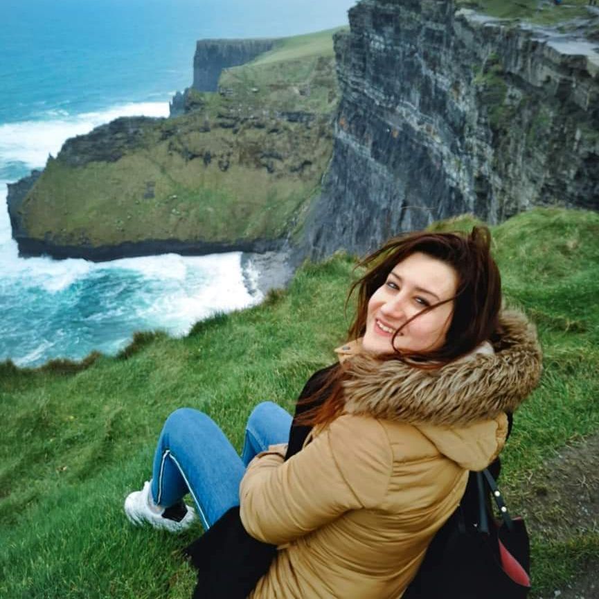 Una donna sorridente con un cappotto invernale siede sul bordo di una scogliera erbosa, affacciata su un mare agitato e formazioni rocciose costiere spettacolari.