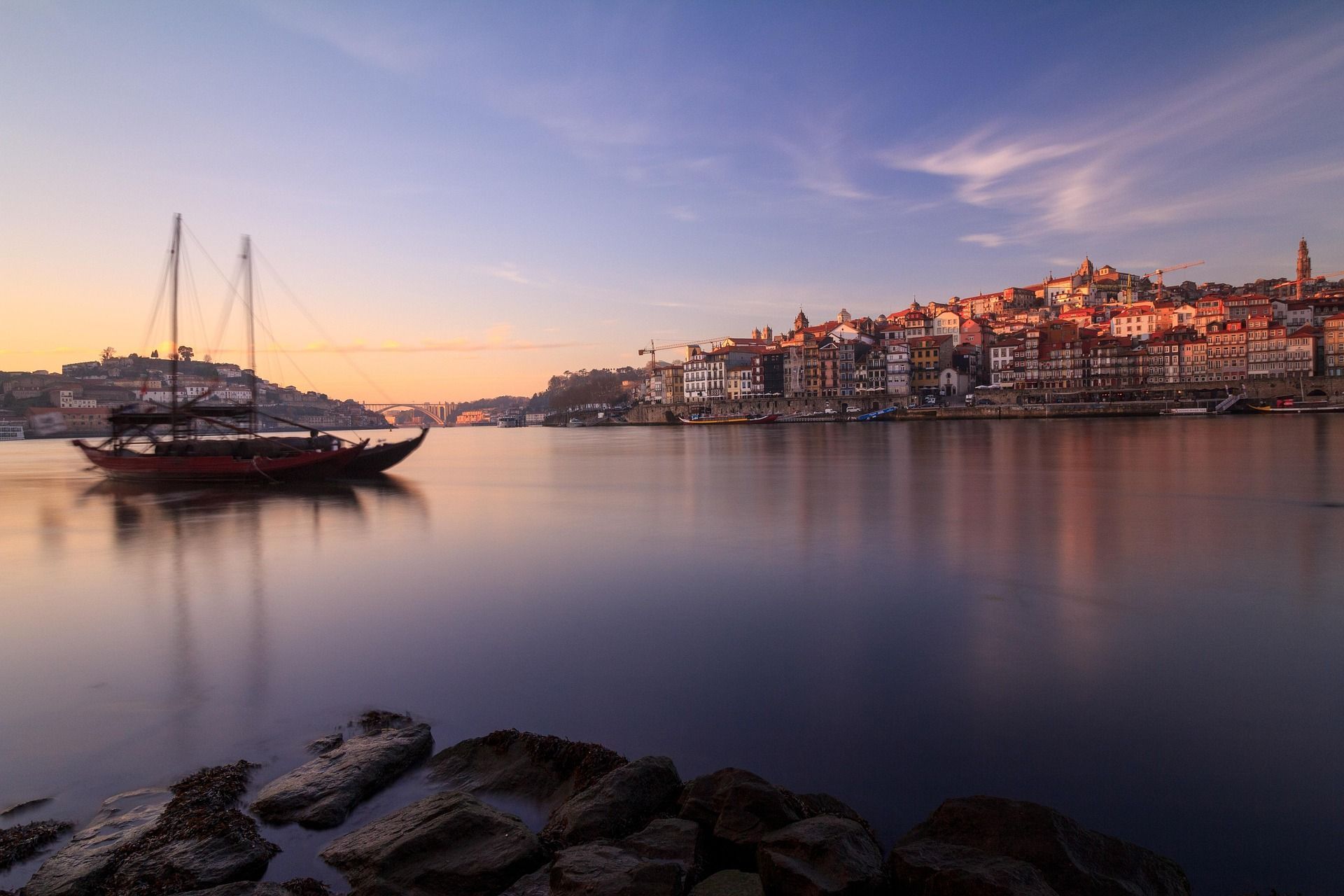 Vista de larga exposición de un barco tradicional en un río tranquilo, frente a una ciudad histórica en una colina bajo un colorido cielo al atardecer.
