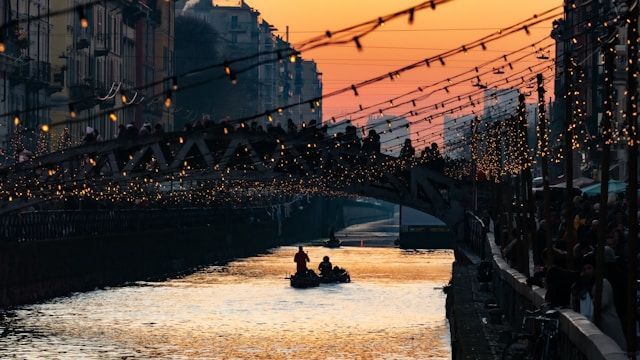 A small boat with people paddles on a canal under a crowded bridge decorated with festive lights during a warm sunset.
