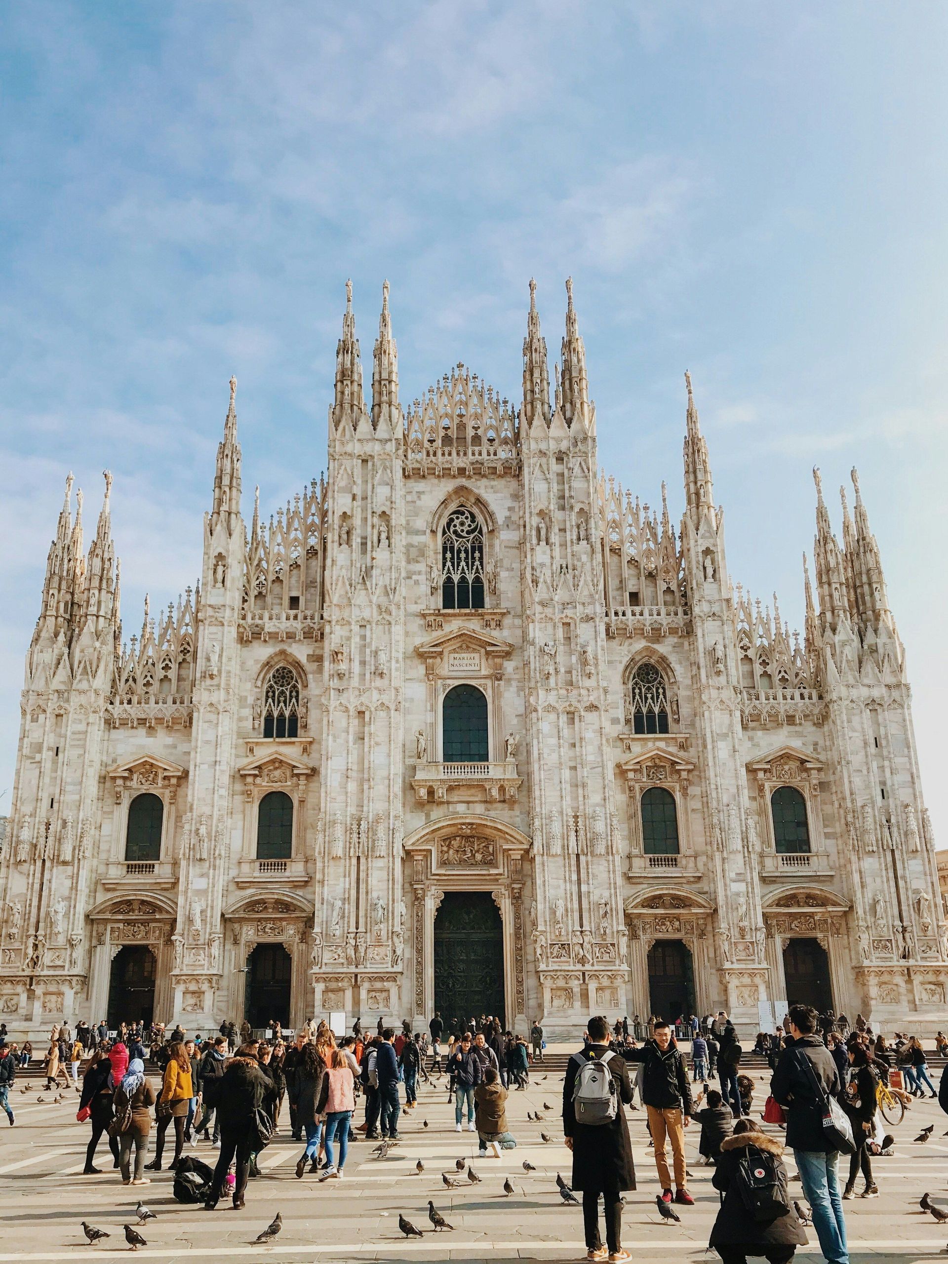 The ornate, spired facade of a large gothic cathedral with a crowd of people and pigeons gathered in the square below.