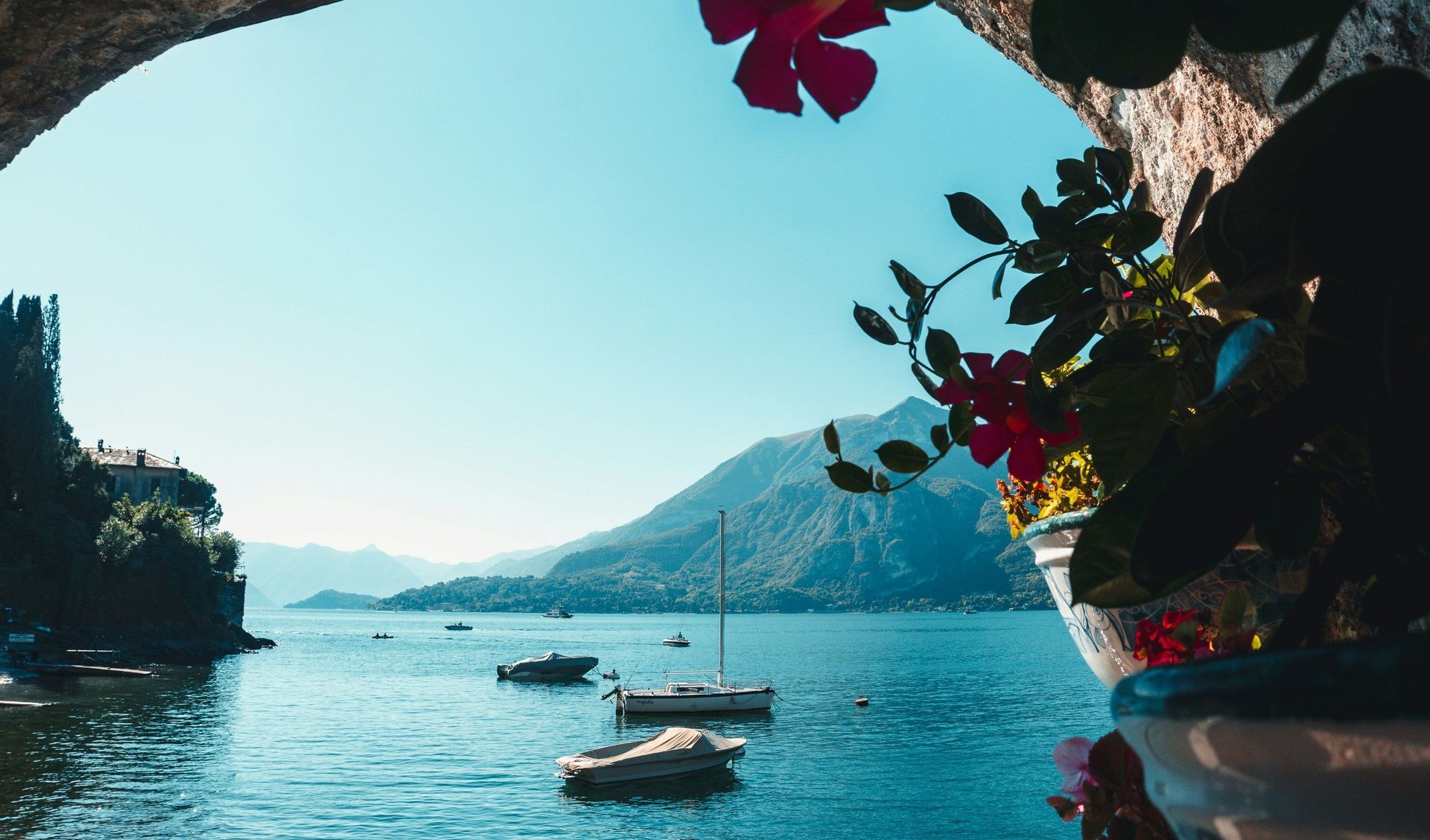 A view of a large lake with several boats, seen from under a stone archway with pink flowers in the foreground and mountains in the background.