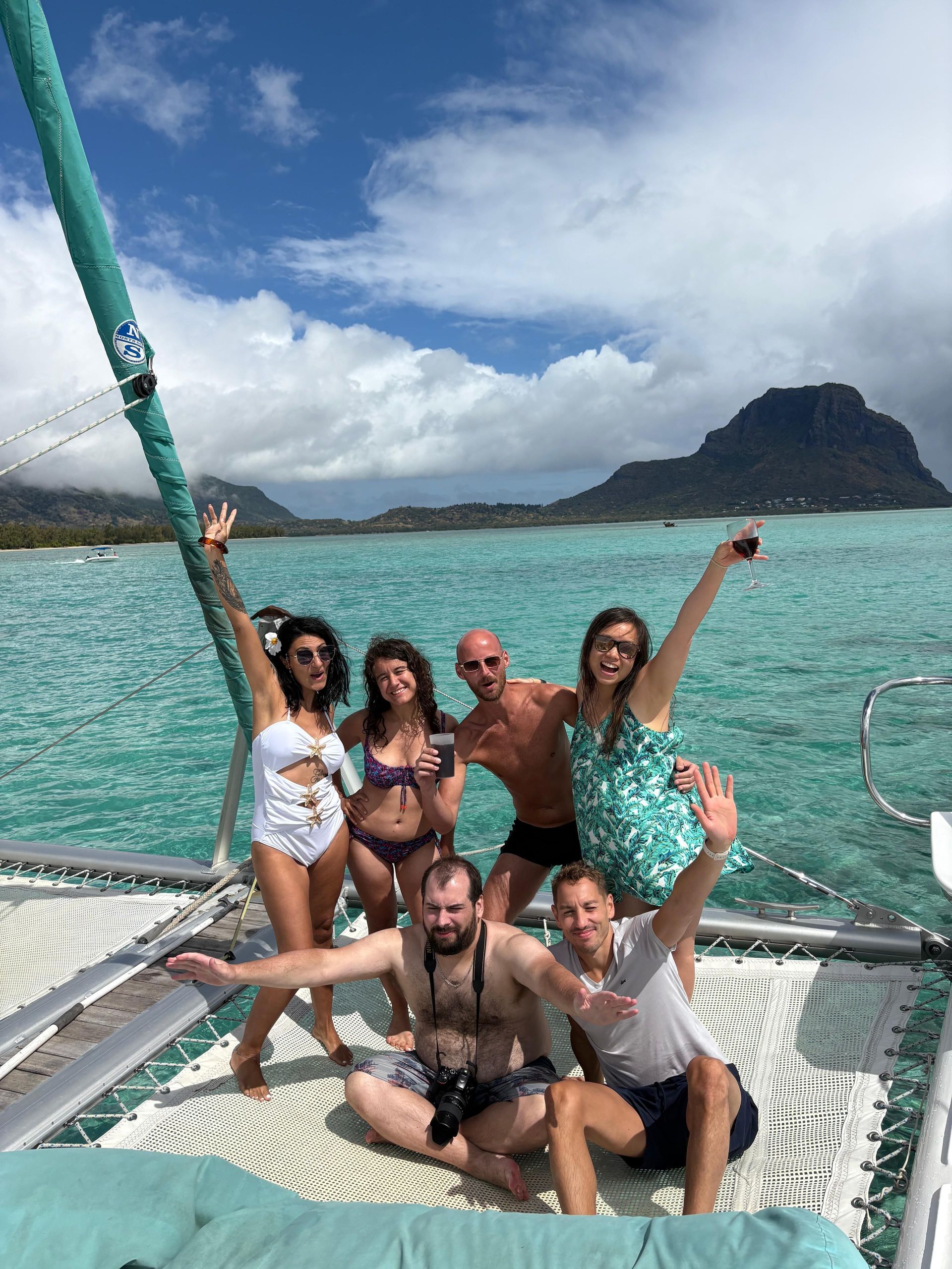Un groupe WeRoad de six personnes posant pour une photo sur le filet d'un catamaran, naviguant sur des eaux turquoise près de montagnes.