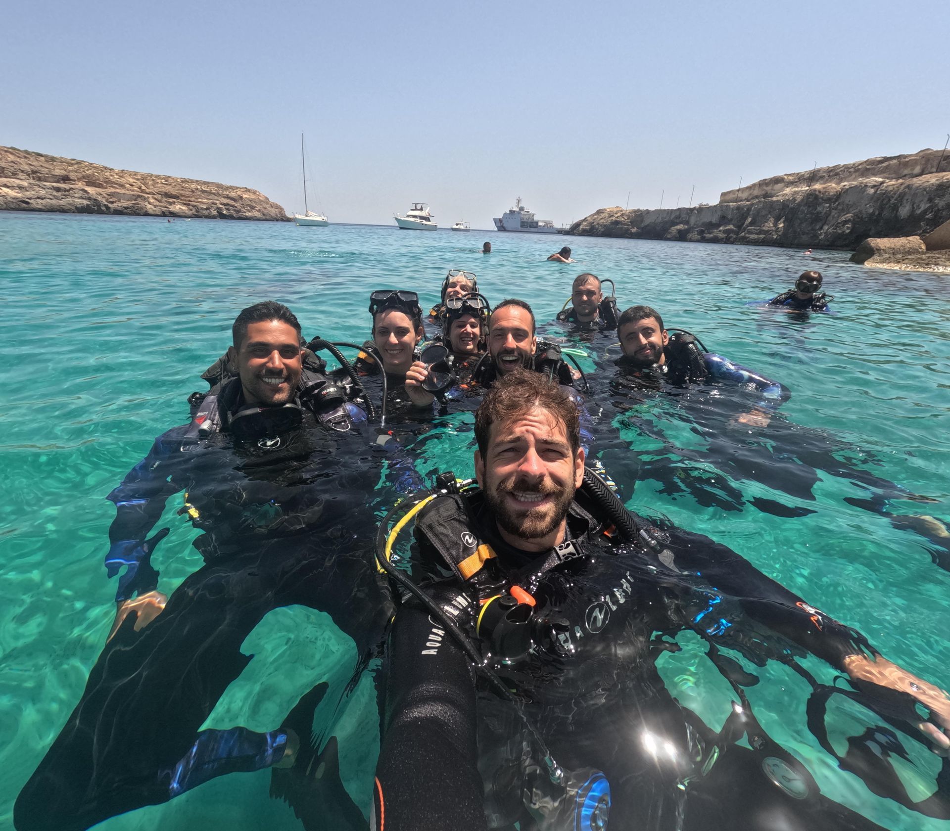 Un gruppo WeRoad in gita subacquea sorride per un selfie mentre galleggia in acque limpide e turchesi vicino a una costa rocciosa.