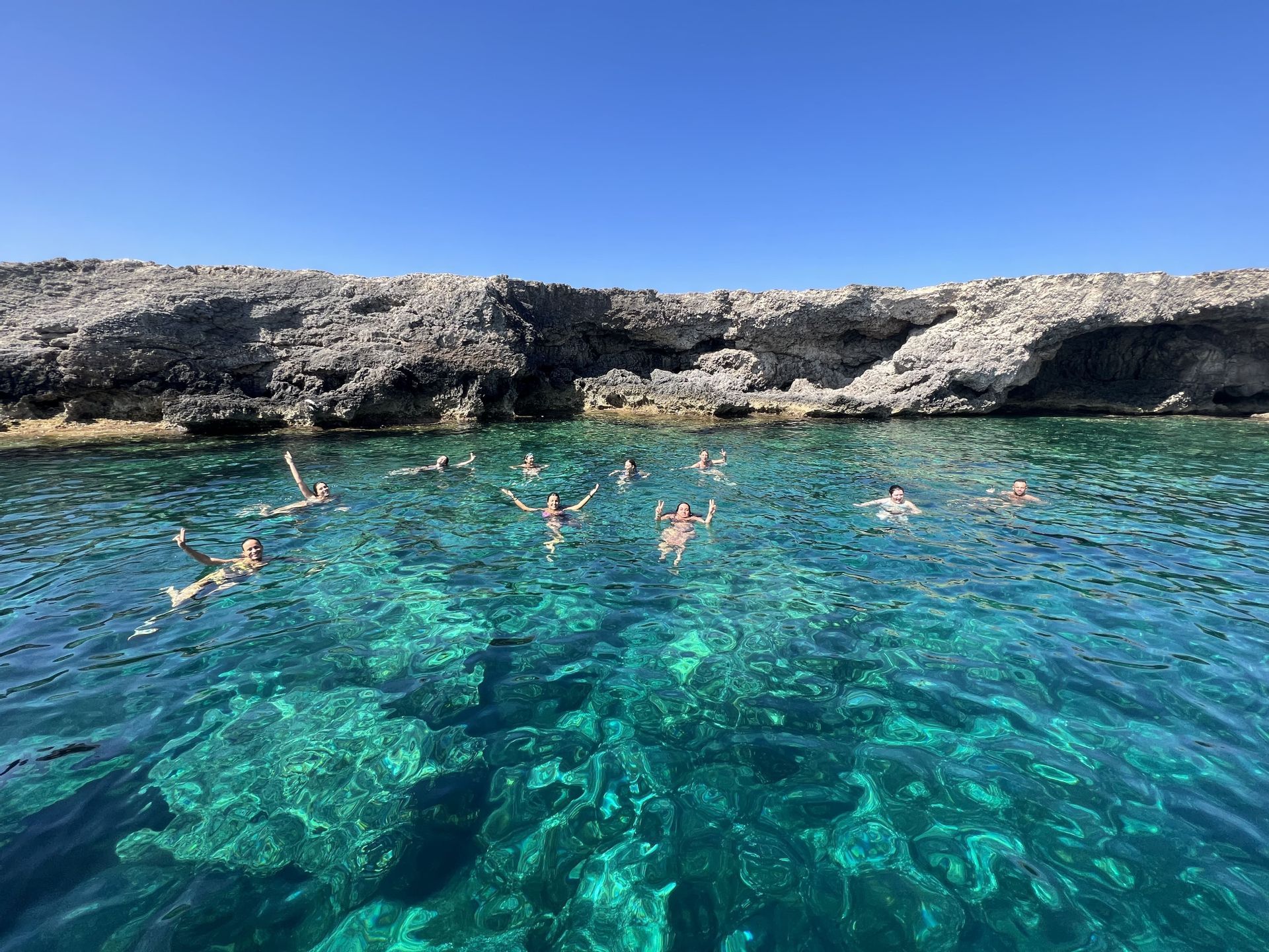 Un viaggio di gruppo WeRoad fa il bagno in acque limpide e turchesi vicino a una costa rocciosa sotto un cielo azzurro.
