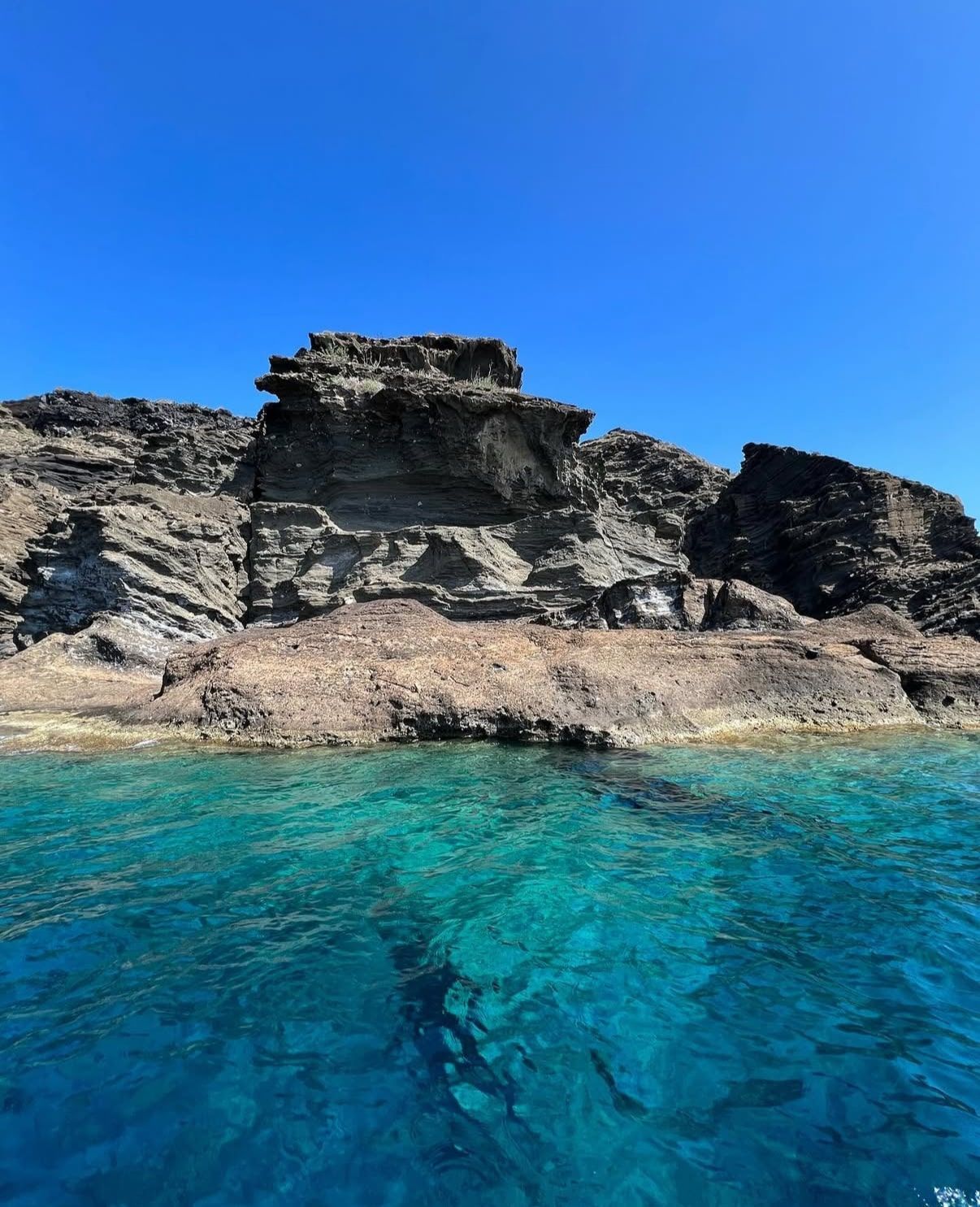 Una scogliera di roccia stratificata si erge da acque marine turchesi e limpide sotto un cielo blu luminoso e senza nuvole.