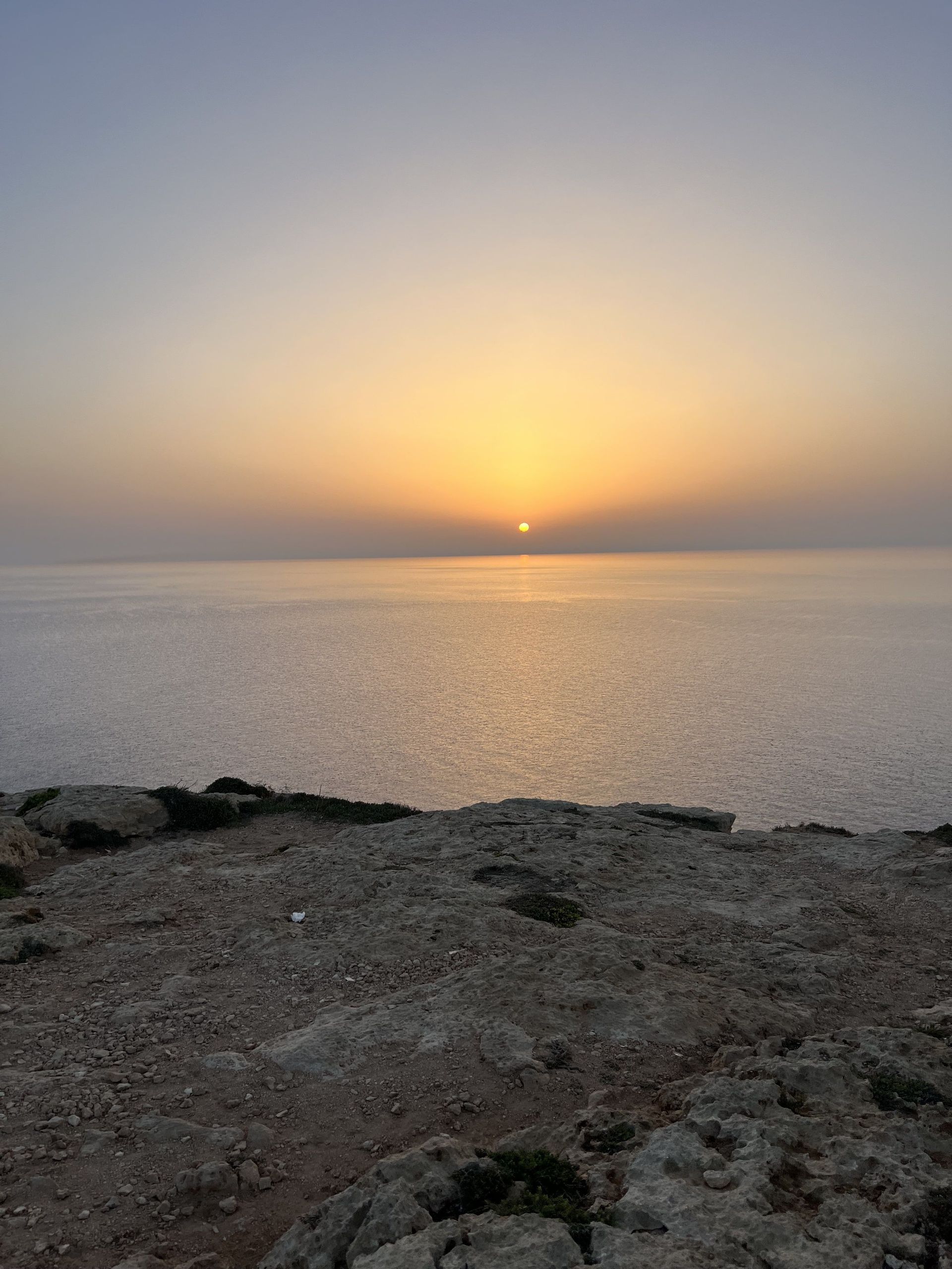 Il sole tramonta all'orizzonte su un mare calmo, visto da una scogliera rocciosa in primo piano.