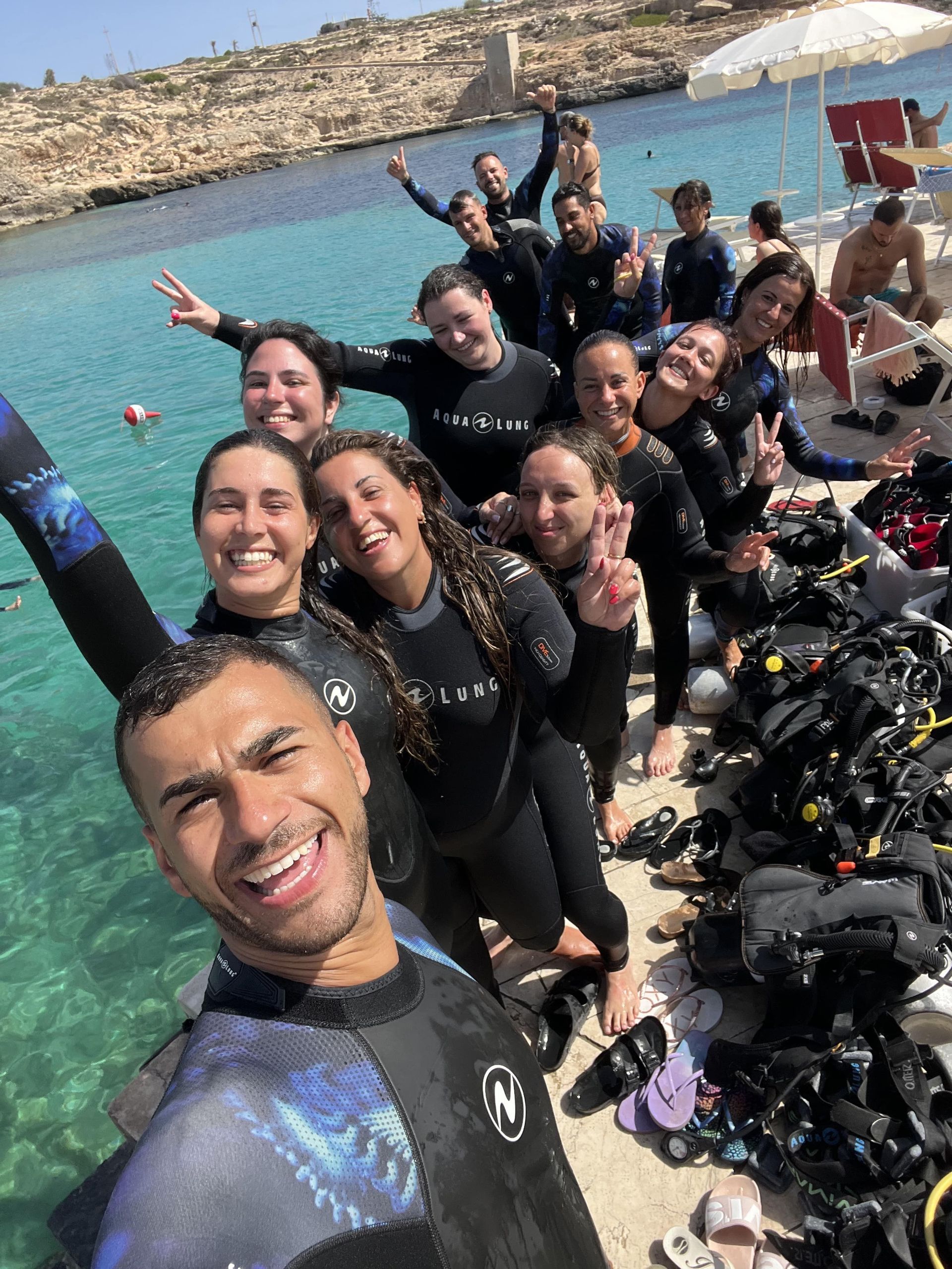 Un viaggio di gruppo WeRoad in mute sorride per un selfie di gruppo su un pontile vicino al mare, con l'attrezzatura da sub pronta.