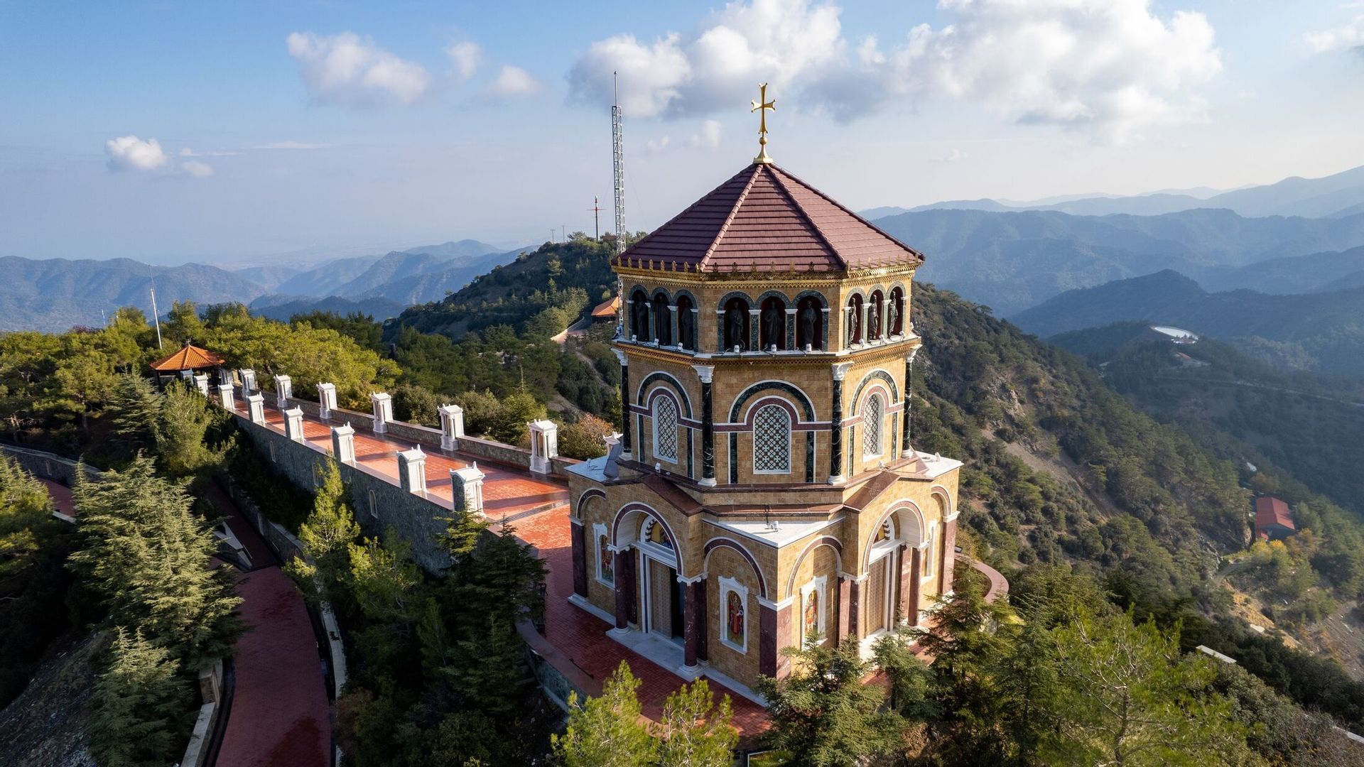 Une église ornée d'un toit de tuiles rouges est perchée sur un sommet boisé, dominant une vaste chaîne de montagnes sous un ciel partiellement nuageux.