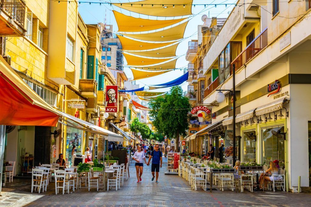 Un couple se promène dans une rue piétonne animée bordée de cafés et de boutiques, sous de grands stores de soleil jaunes suspendus entre les bâtiments.