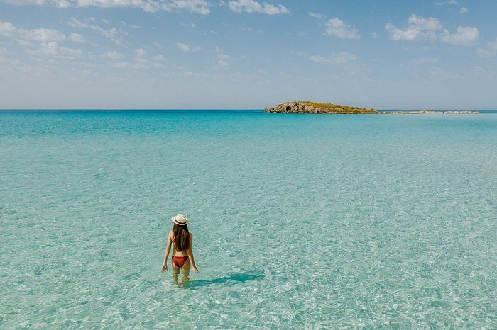 Une femme portant un chapeau de soleil et un bikini se tient dans une eau turquoise peu profonde et limpide, regardant vers une petite île à l'horizon.