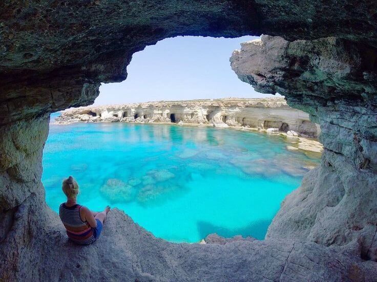 Une femme est assise à l'entrée d'une grotte marine, contemplant une baie calme aux eaux turquoise claires et des falaises rocheuses sous un ciel bleu.