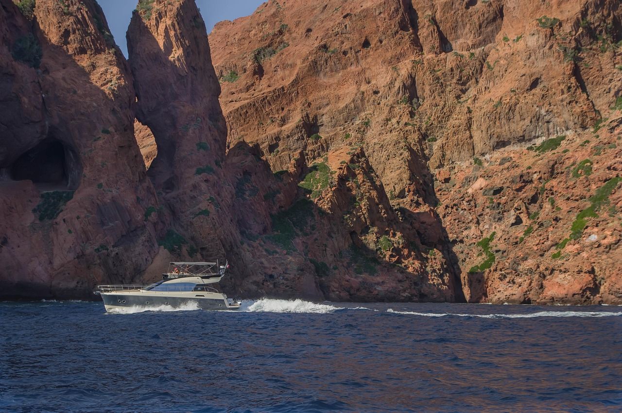 Un yacht à moteur navigue sur la mer d'un bleu profond le long d'une côte spectaculaire de falaises de roche rouge avec une arche naturelle.