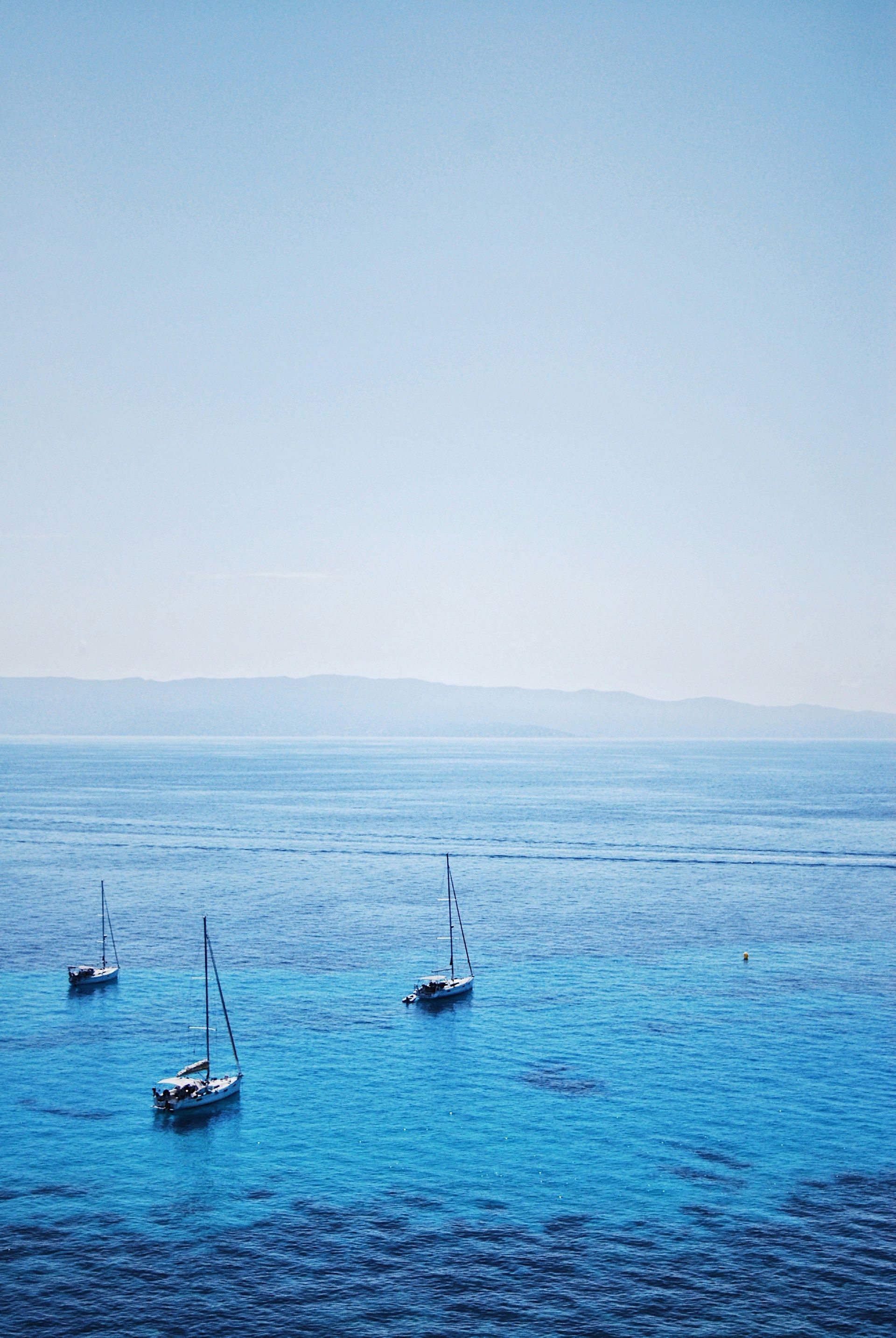 Trois voiliers flottent sur une eau calme et turquoise près d'un rivage lointain sous un ciel clair.