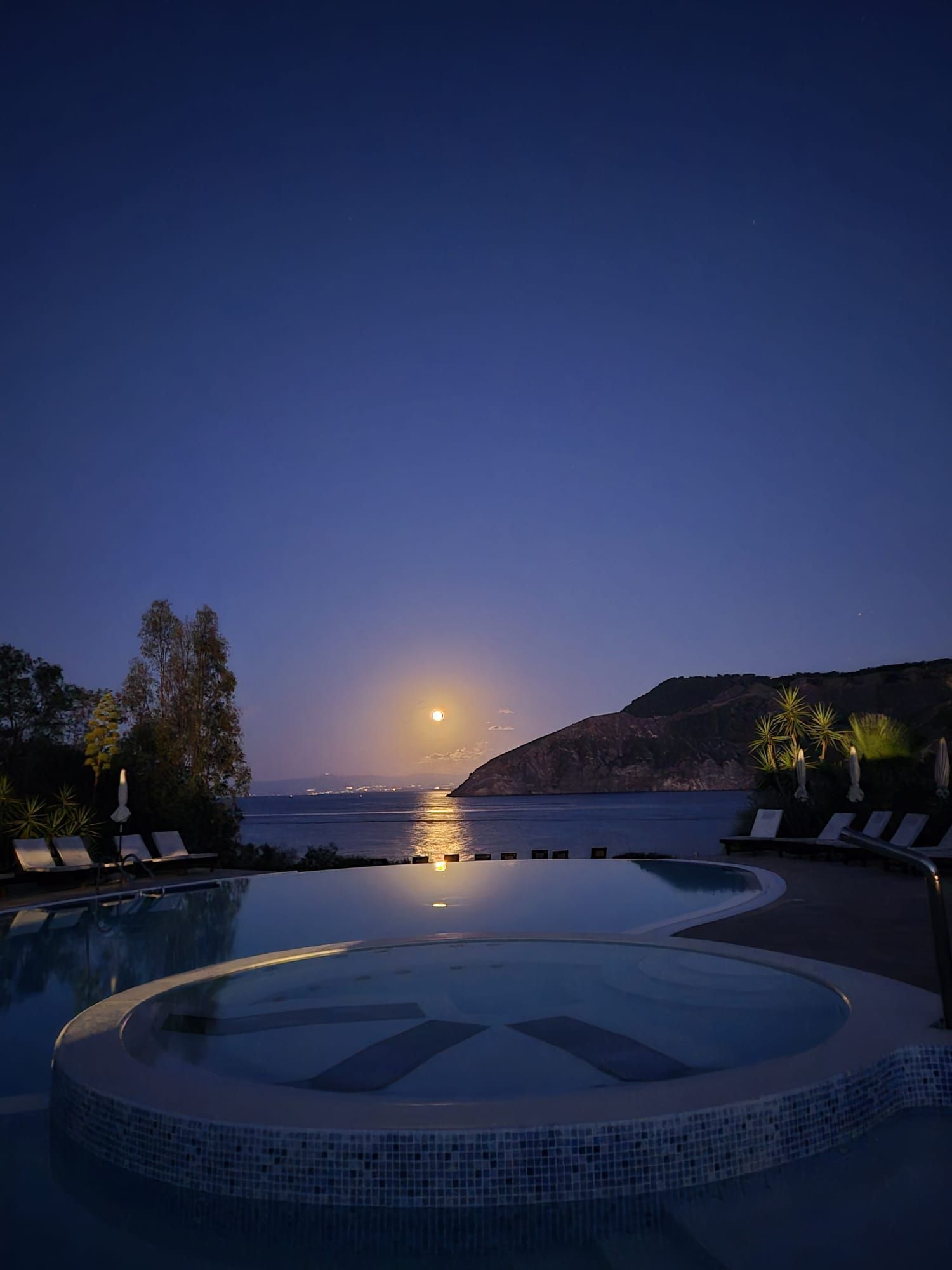 Una piscina di notte con vista sulla luna piena che si riflette sul mare calmo, vicino a una costa scura.