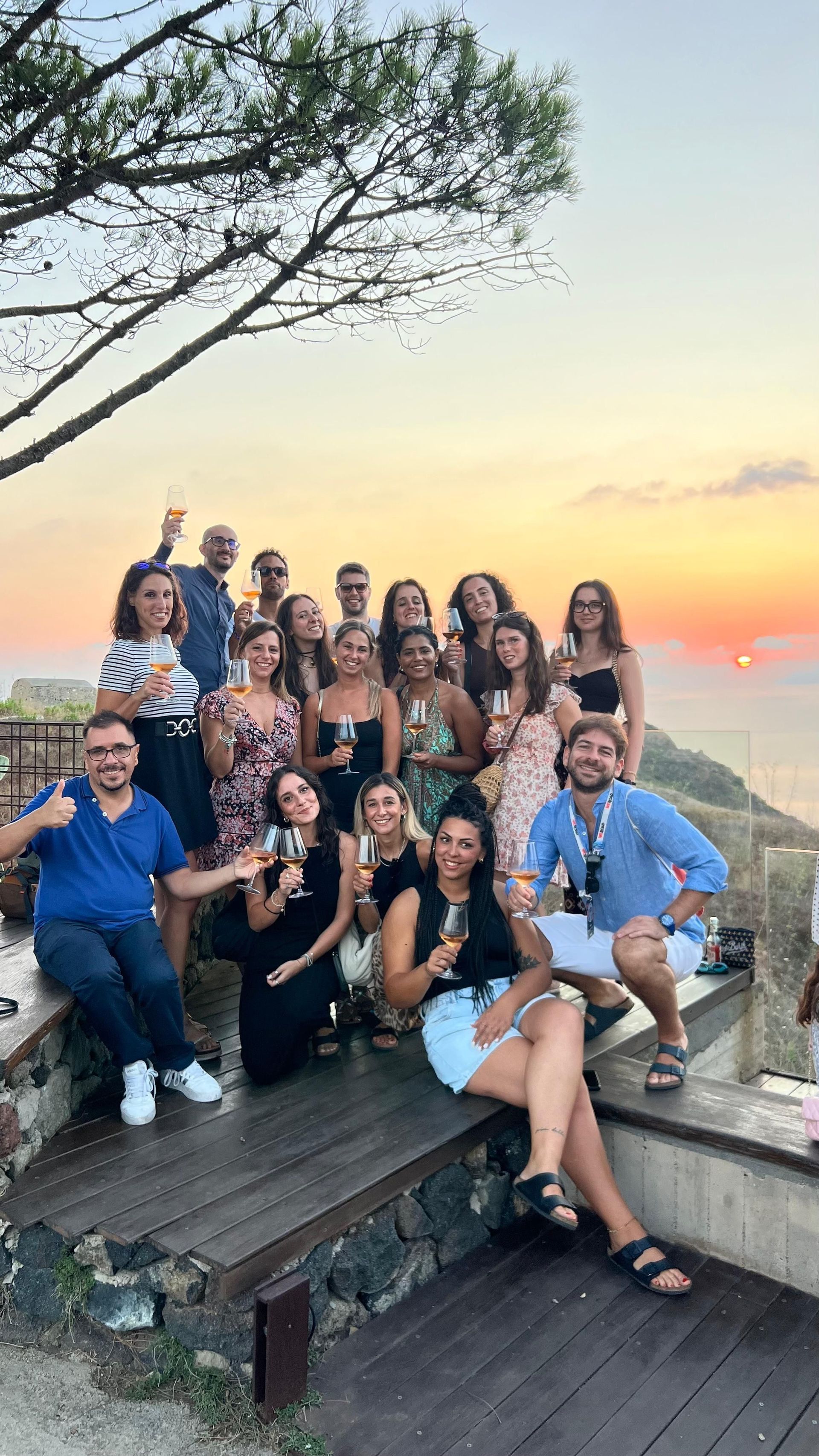 Un gruppo WeRoad in posa per una foto su una terrazza, con calici di vino, durante un tramonto dorato sul mare.