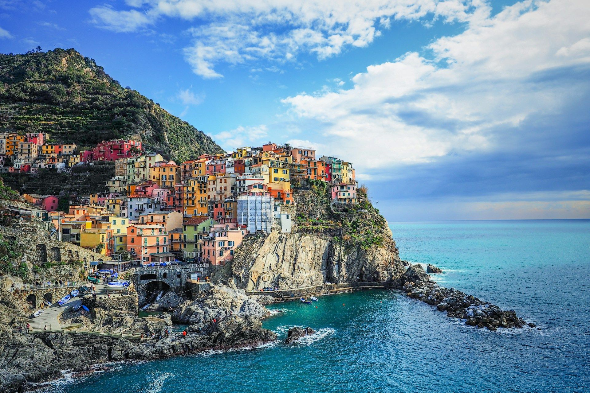 A village of colorful houses built on a rocky cliffside, overlooking a calm blue sea under a partly cloudy sky.
