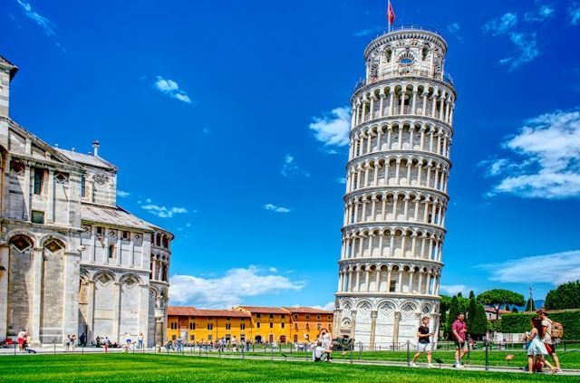 The Leaning Tower of Pisa stands on a green lawn next to a large cathedral under a bright blue sky, with people strolling nearby.