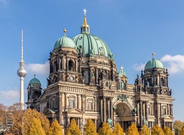 Une cathédrale ornée avec de grands dômes verts et une haute tour de télévision en arrière-plan, vue au-dessus d'arbres d'automne jaunes.