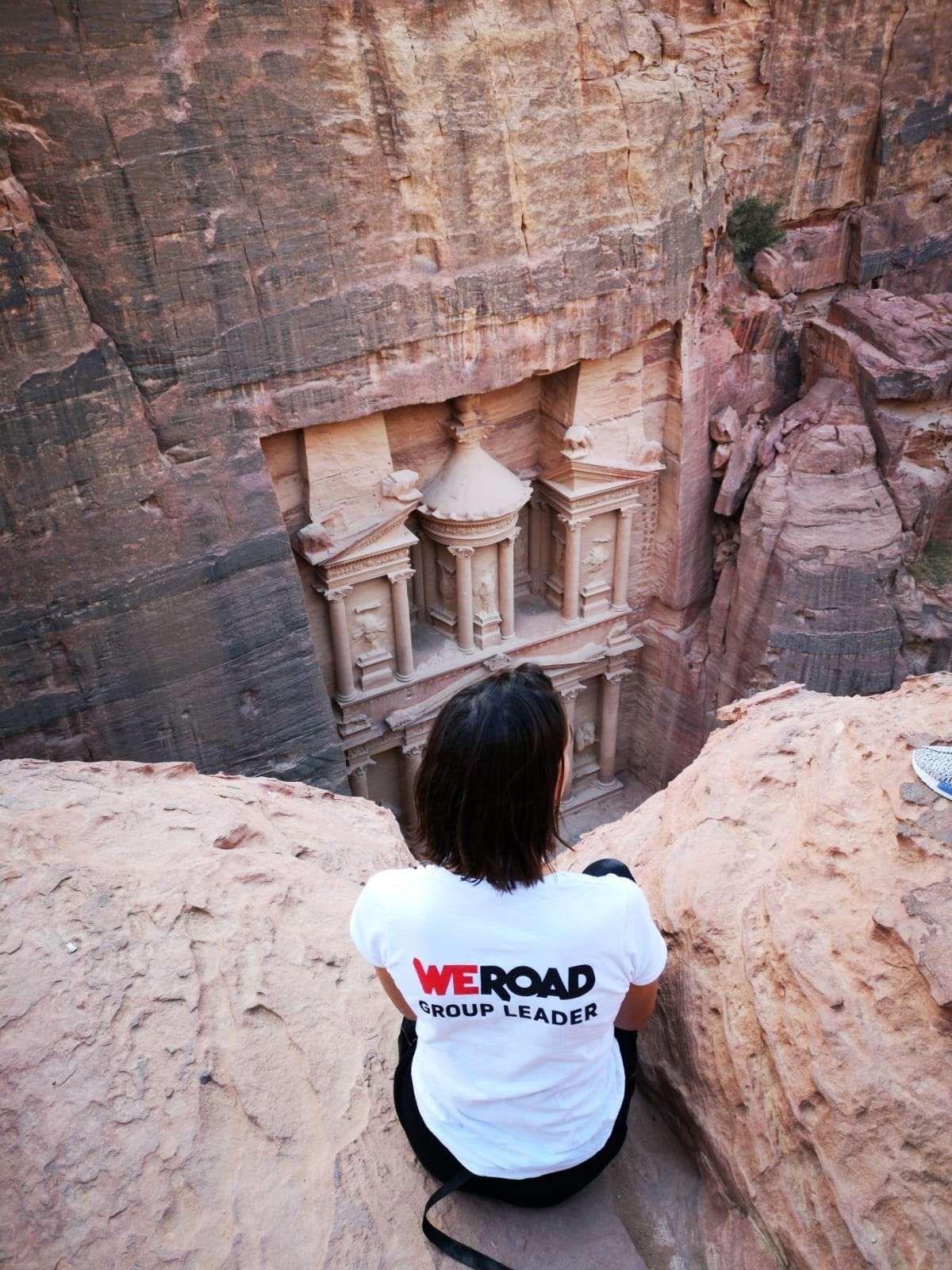 Un Group Leader WeRoad, di spalle, siede sul bordo di una scogliera rocciosa, ammirando un tempio scavato in un canyon di roccia rossa.