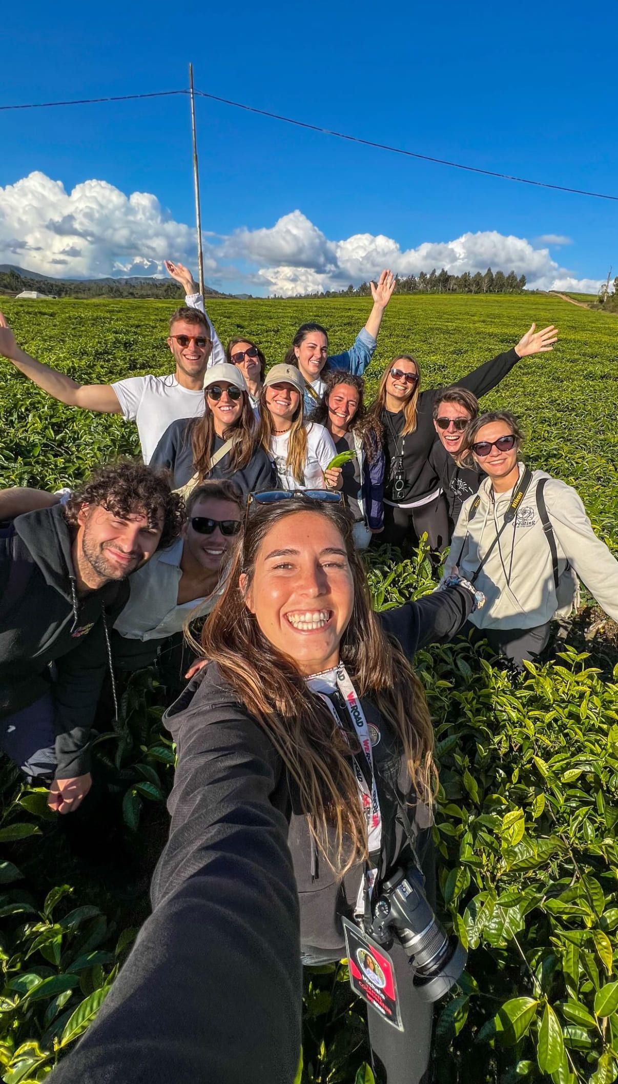 Un gruppo WeRoad in viaggio sorride per un selfie in una piantagione di tè verde, sotto un cielo azzurro e nuvole.