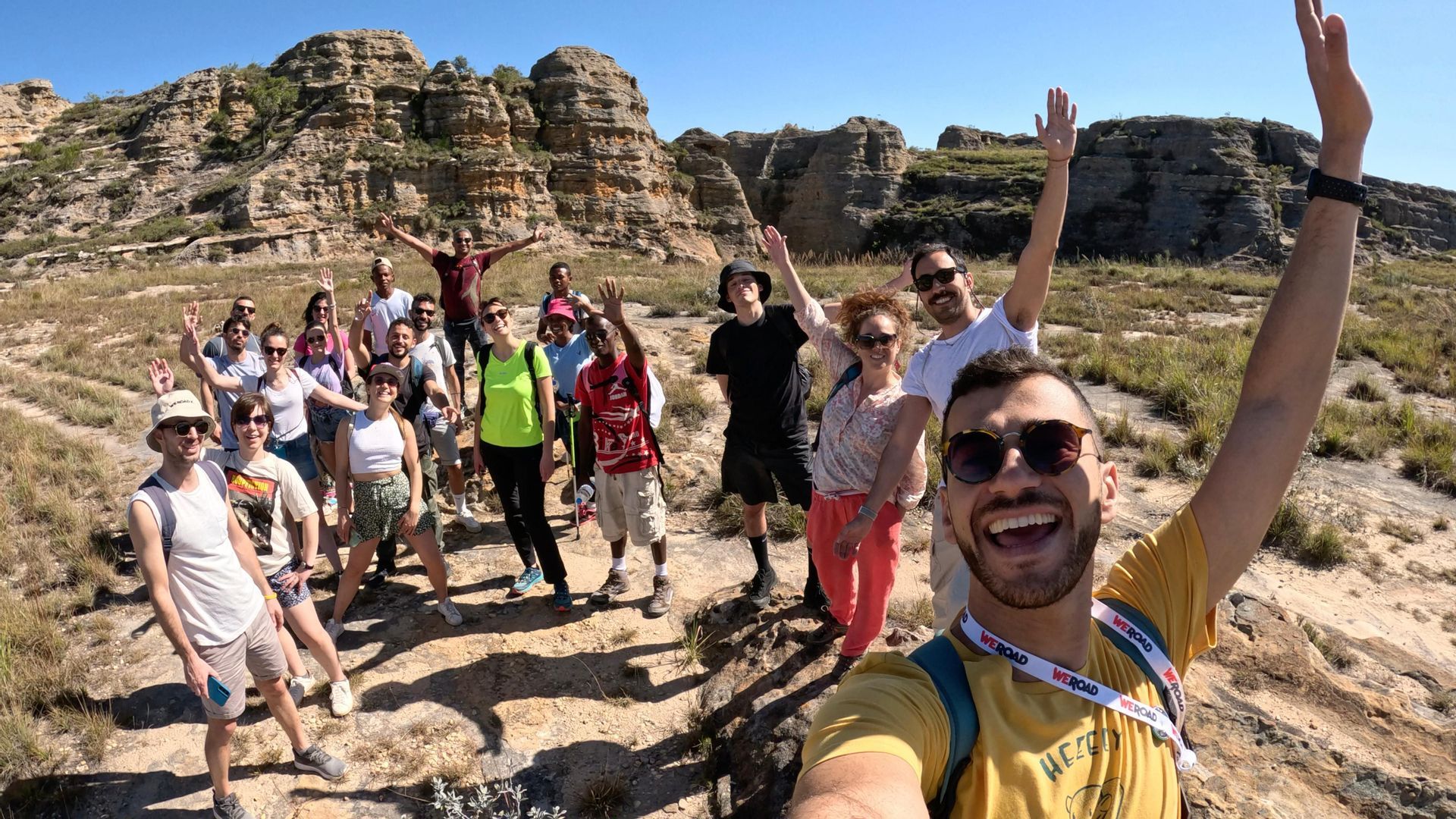 Un gruppo WeRoad si scatta un selfie durante un'escursione in un paesaggio roccioso e soleggiato, con tutti sorridenti e che salutano.