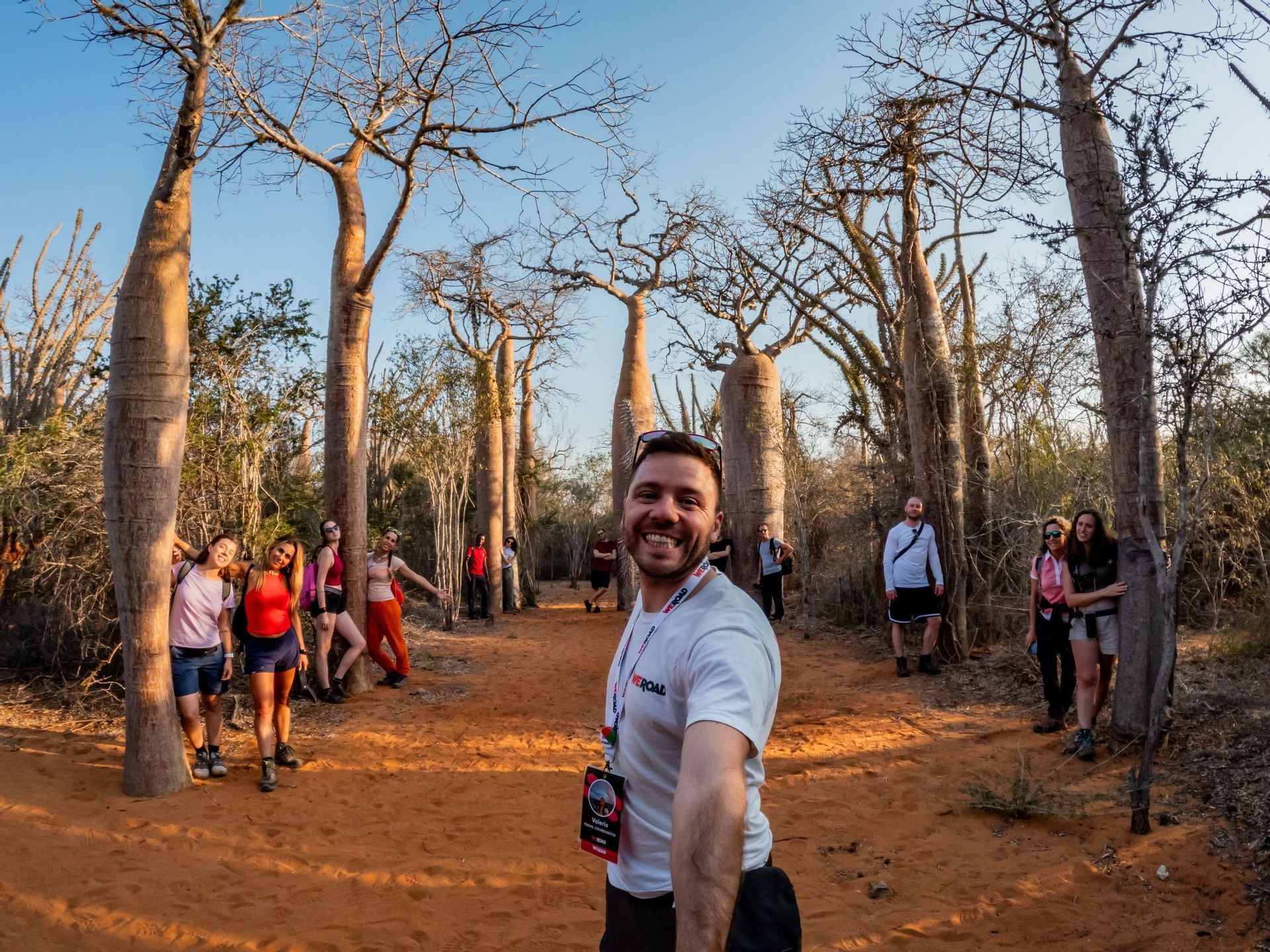 Un uomo sorridente si scatta un selfie con il suo gruppo WeRoad tra grandi baobab su un sentiero sterrato.