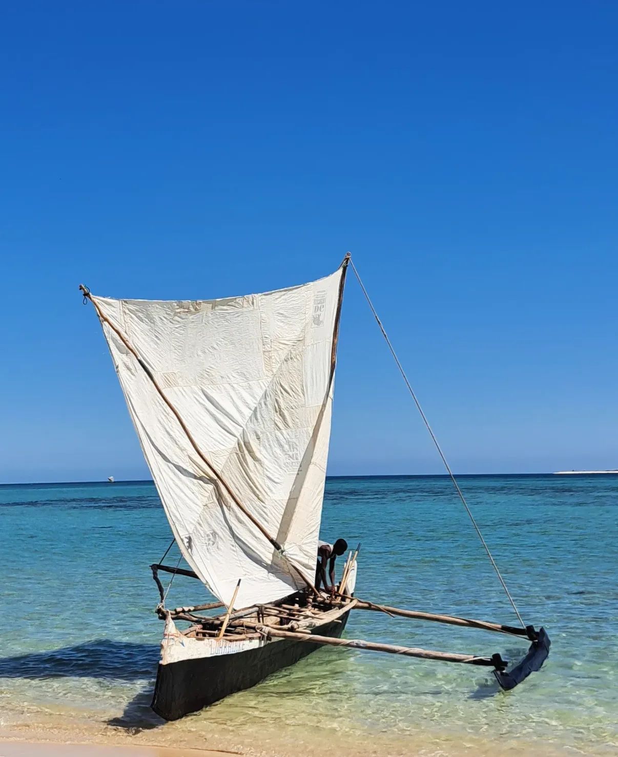 Una tradizionale barca a vela a bilanciere in legno con una grande vela bianca riposa in acque turchesi poco profonde vicino a una riva sabbiosa.