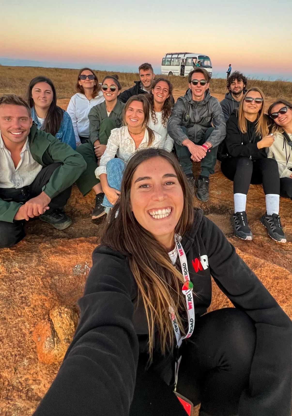 Una donna si scatta un selfie con il suo gruppo di viaggio WeRoad mentre sono seduti su uno sperone roccioso al tramonto.