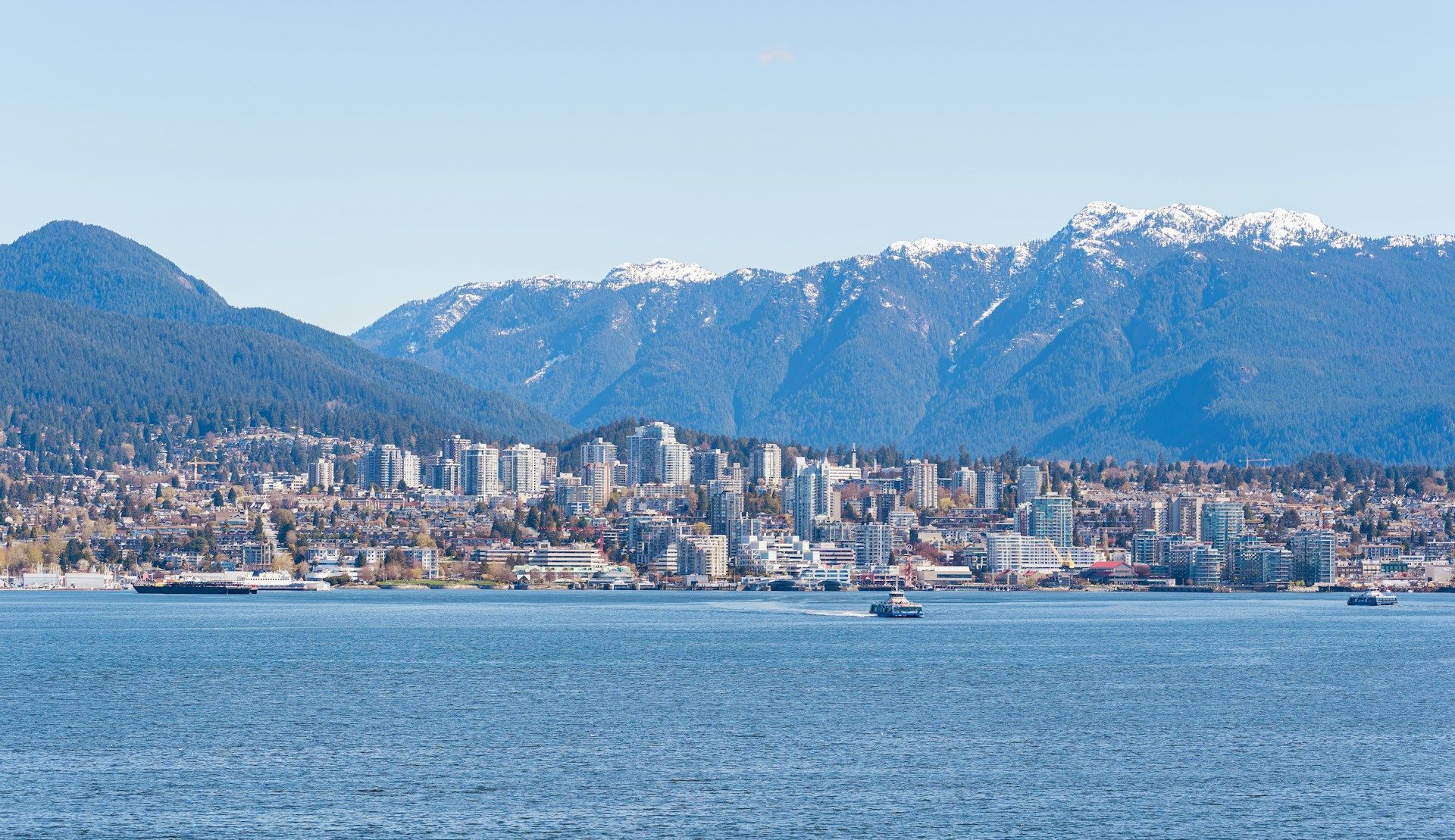 Uno skyline cittadino su uno specchio d'acqua, con grandi montagne innevate che si ergono sullo sfondo sotto un cielo sereno.