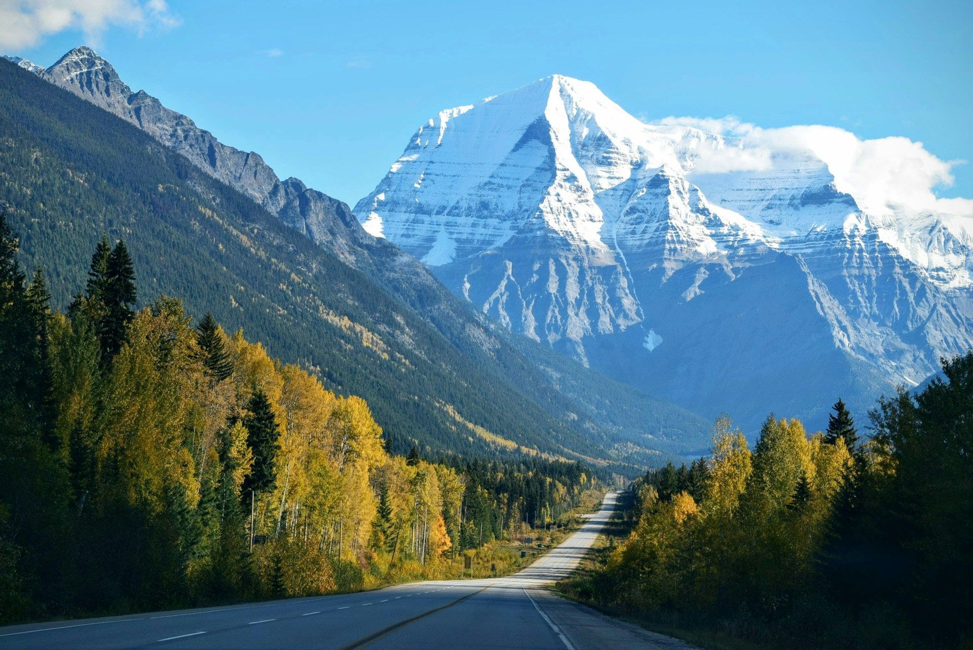 Una strada asfaltata attraversa una valle fiancheggiata da alberi autunnali verso un'imponente montagna innevata sotto un cielo azzurro.