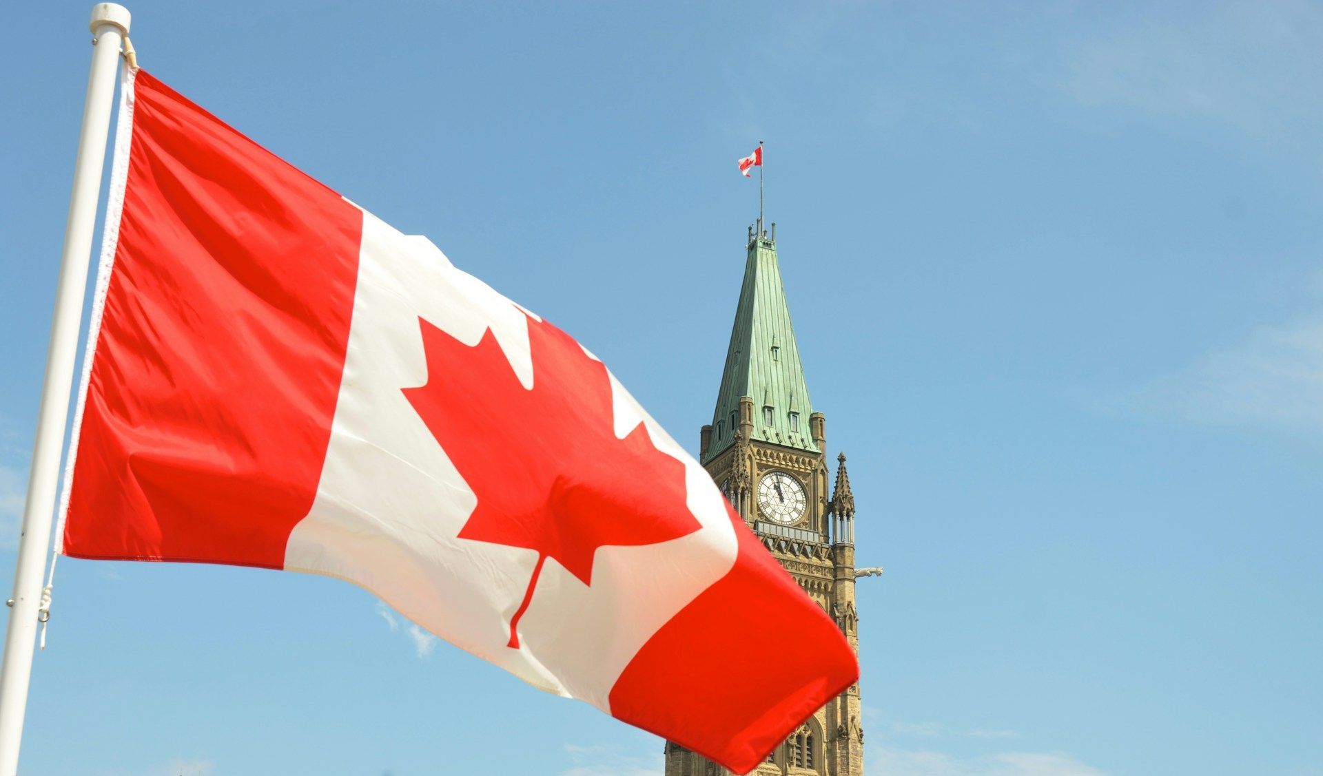 La bandiera canadese sventola su un pennone davanti a una torre dell'orologio in pietra contro un cielo azzurro chiaro.