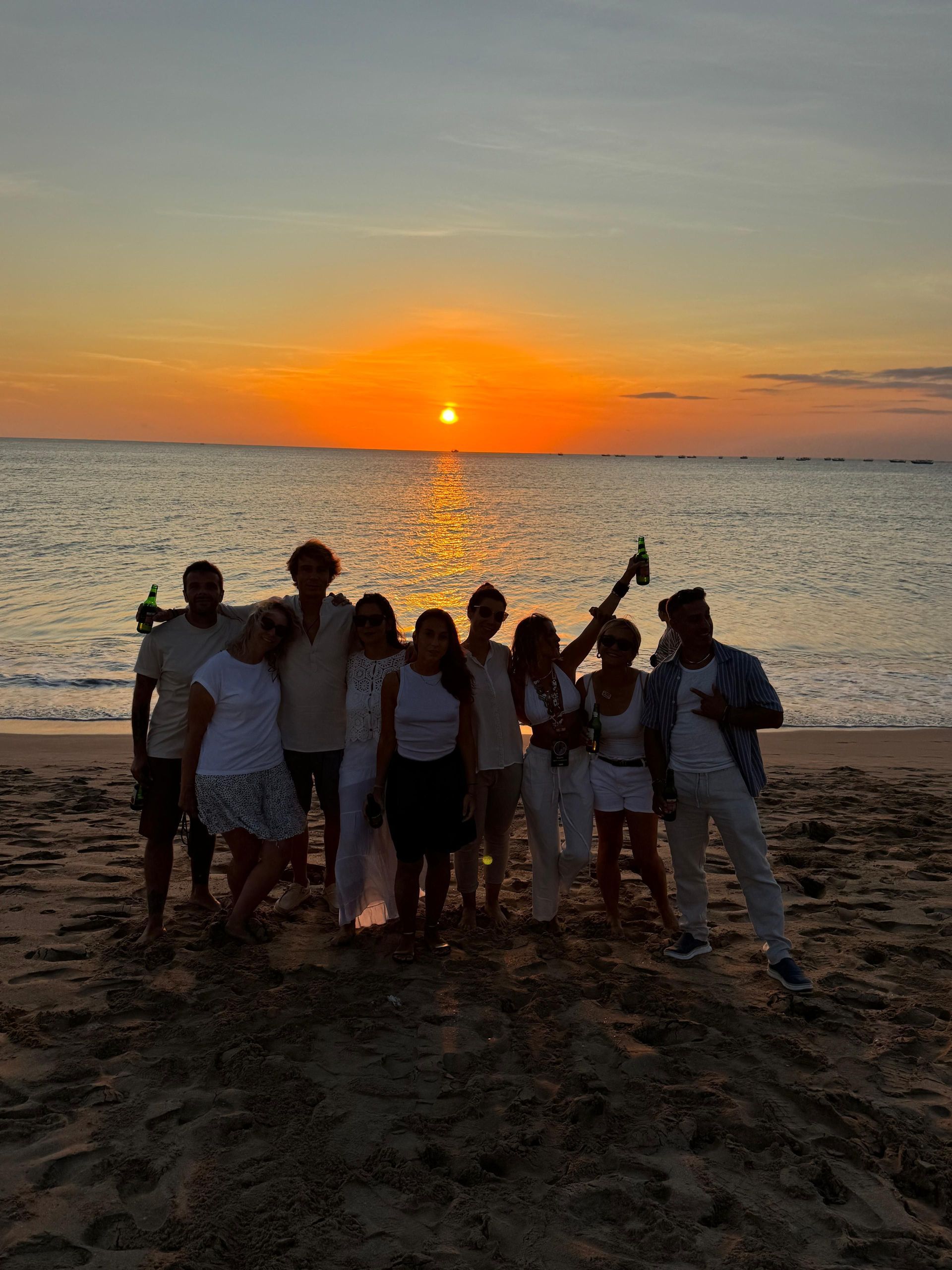 Un gruppo WeRoad posa su una spiaggia sabbiosa, stagliato contro un vibrante tramonto arancione sul mare.