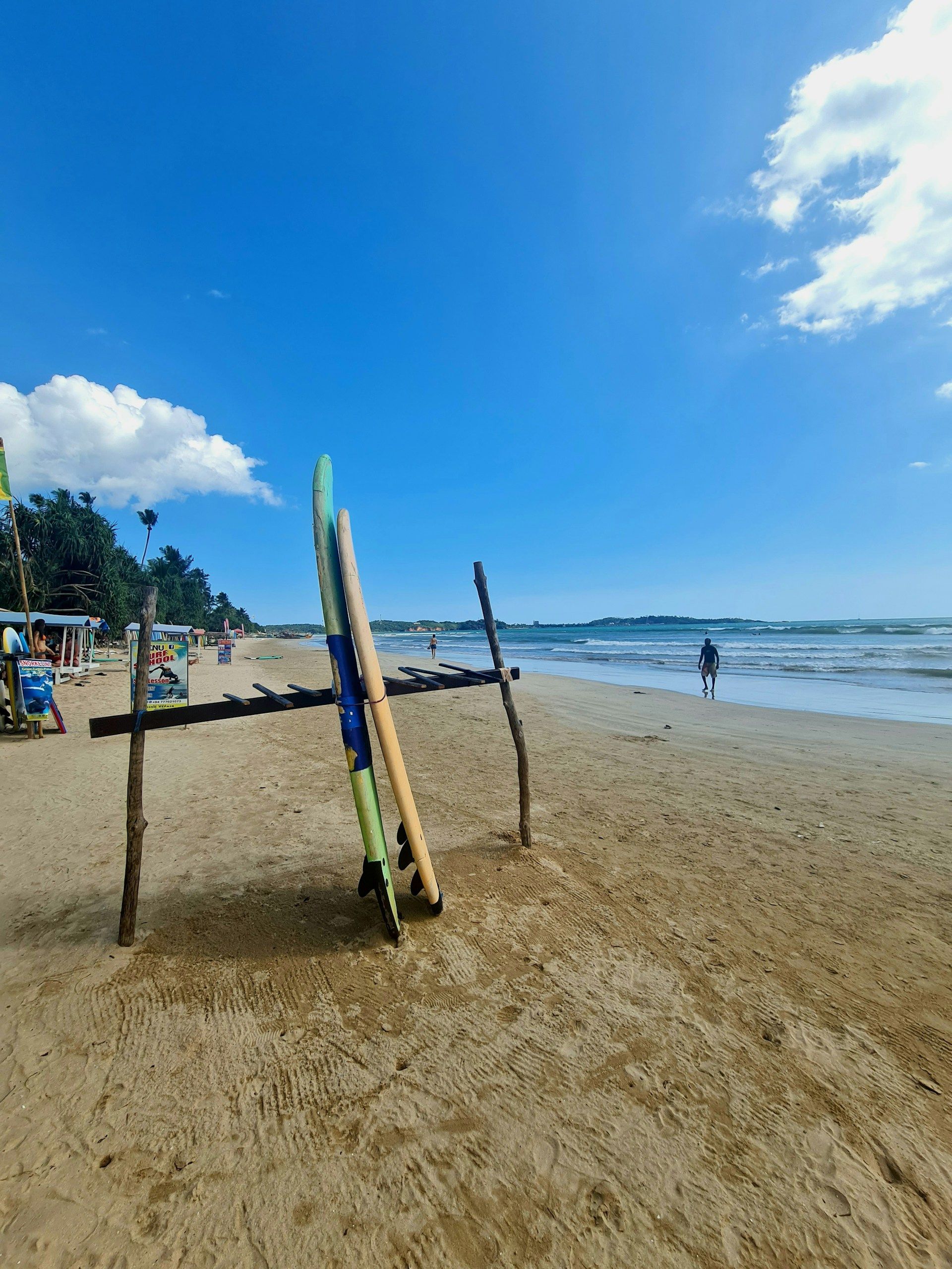 Due tavole da surf appoggiate a un portatavole di legno su una spiaggia sabbiosa, con l'oceano e un cielo azzurro chiaro sullo sfondo.