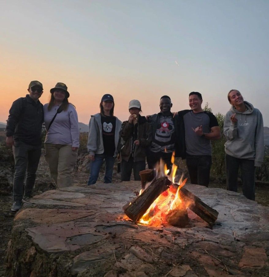 Un voyage en groupe WeRoad avec des gens souriants autour d'un feu de camp sur une colline au coucher du soleil.