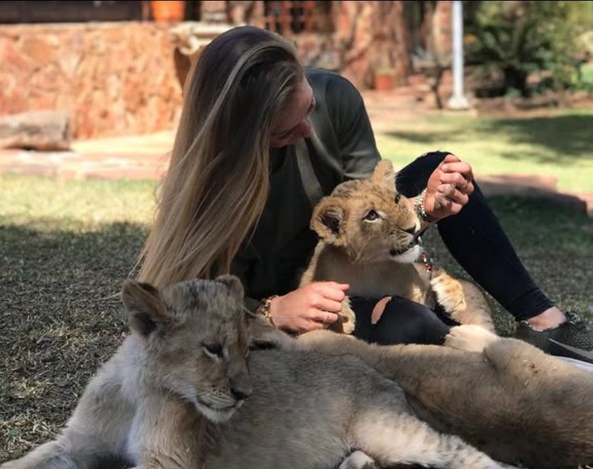 Une femme aux longs cheveux blonds est assise sur une pelouse et caresse deux jeunes lionceaux.