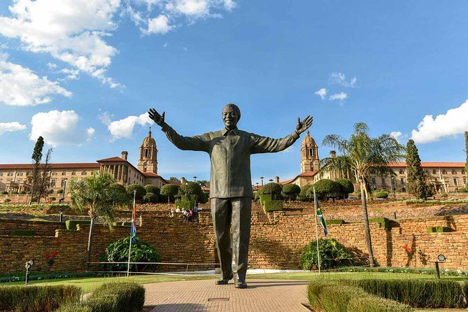 Une grande statue de bronze de Nelson Mandela se dresse, les bras tendus, dans un jardin devant un bâtiment majestueux et symétrique sous un ciel bleu.