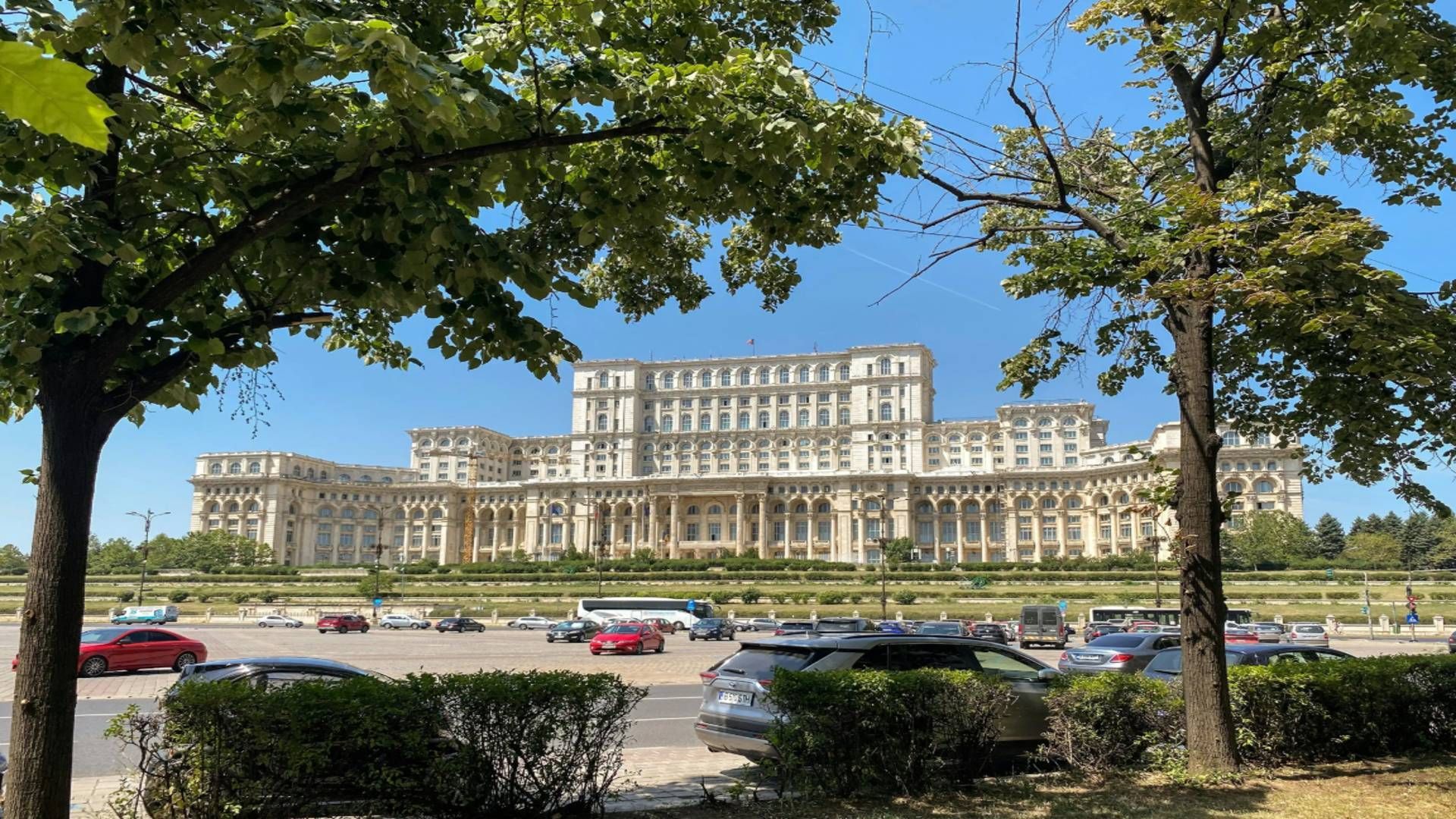 Un maestoso e sfarzoso palazzo, visibile da una strada e un parcheggio, incorniciato da alberi verdi e frondosi sotto un cielo azzurro e limpido.