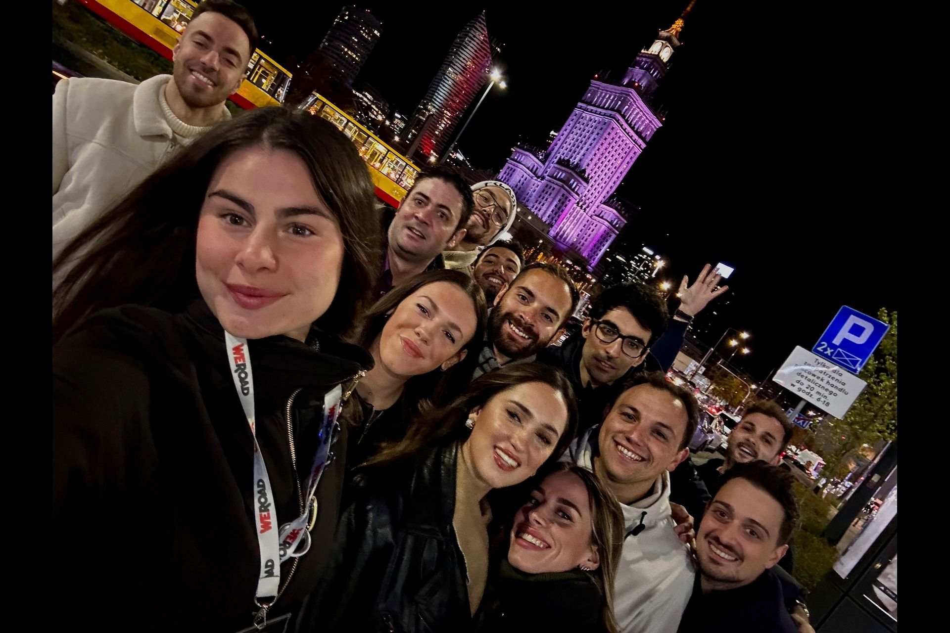 A WeRoad group trip takes a selfie together at night in a city, with an illuminated tower and a tram in the background.