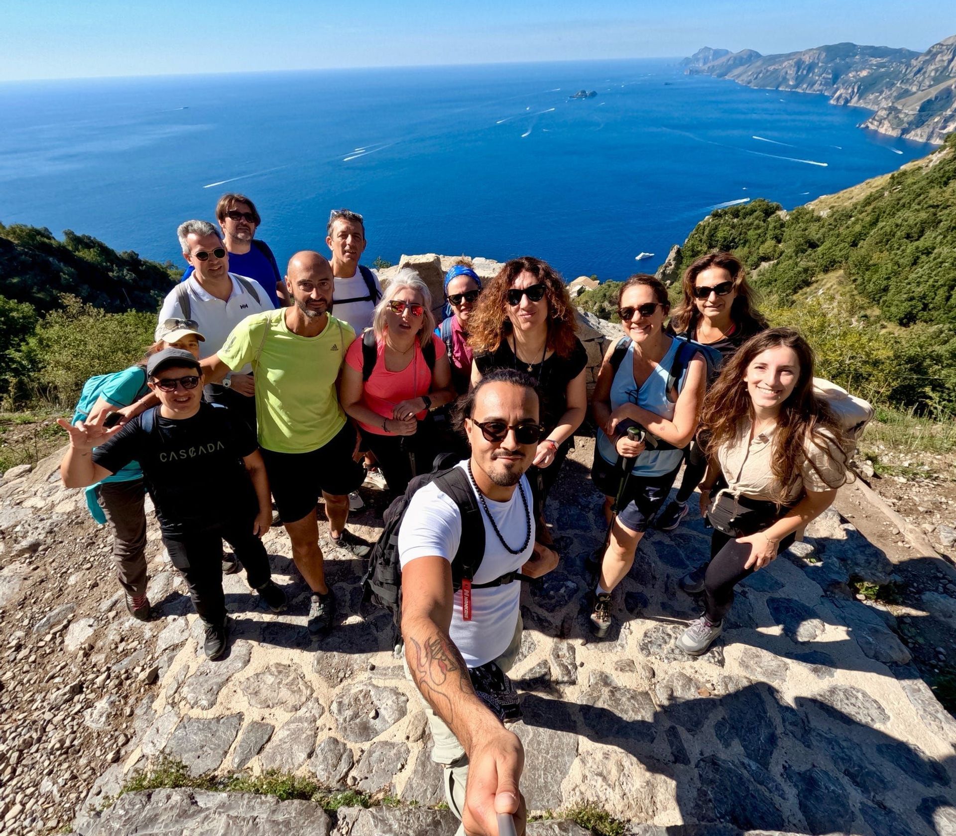 Un gruppo di escursionisti WeRoad si scatta un selfie su una scogliera soleggiata a picco sul mare blu e la costa.