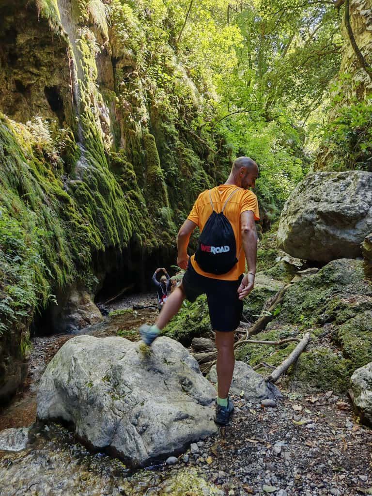 Un uomo con uno zaino WeRoad fa un'escursione in uno stretto canyon muschioso, scavalcando una grande roccia in un ruscello.