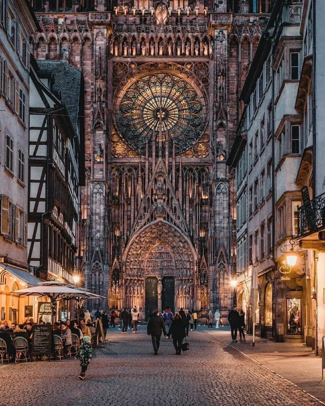 The illuminated facade of a large Gothic cathedral with a detailed rose window, seen from a narrow cobblestone street at dusk.