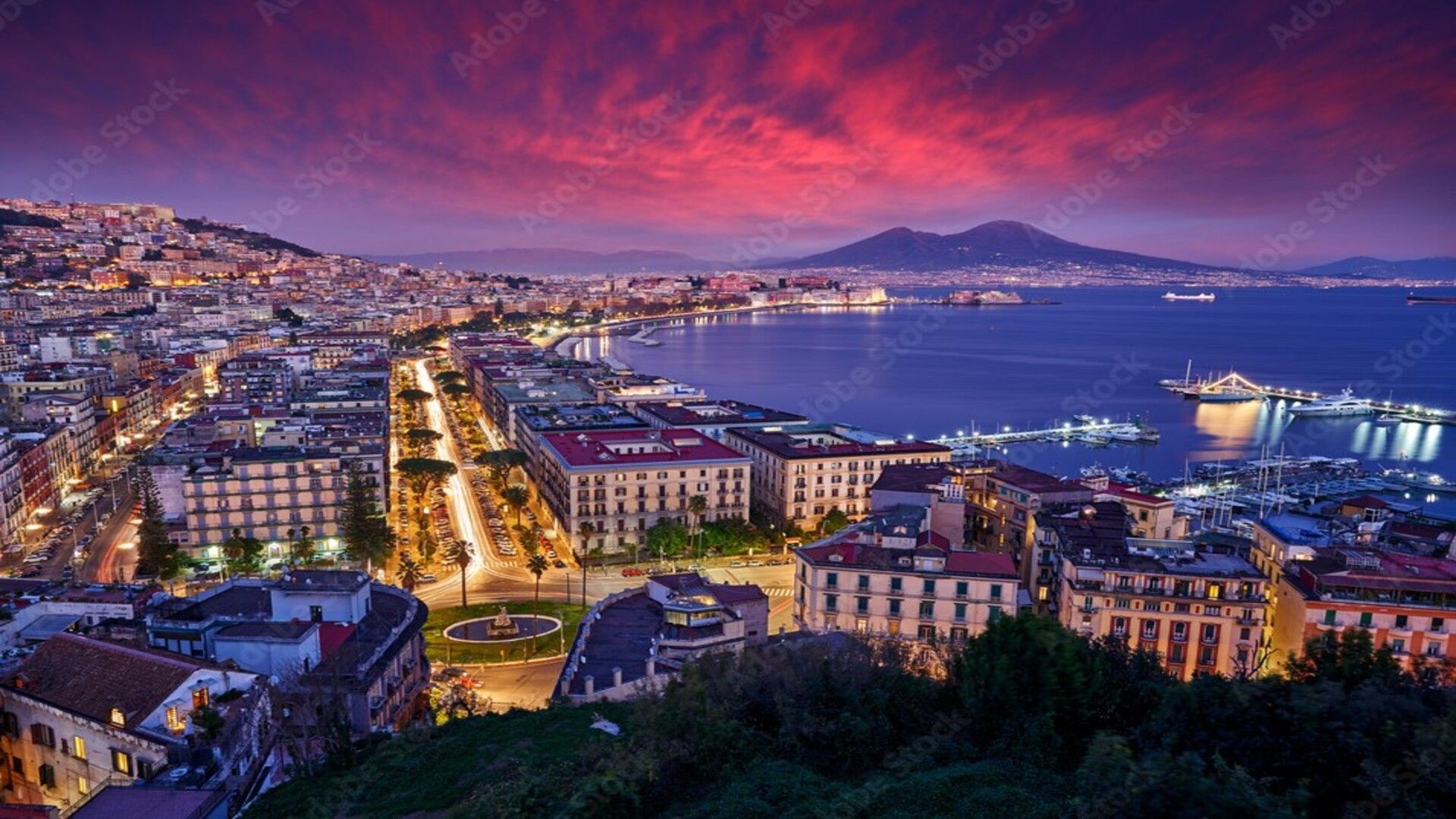 Veduta panoramica di una città costiera illuminata e della sua baia al tramonto, con una grande montagna sullo sfondo sotto un cielo drammatico e colorato.
