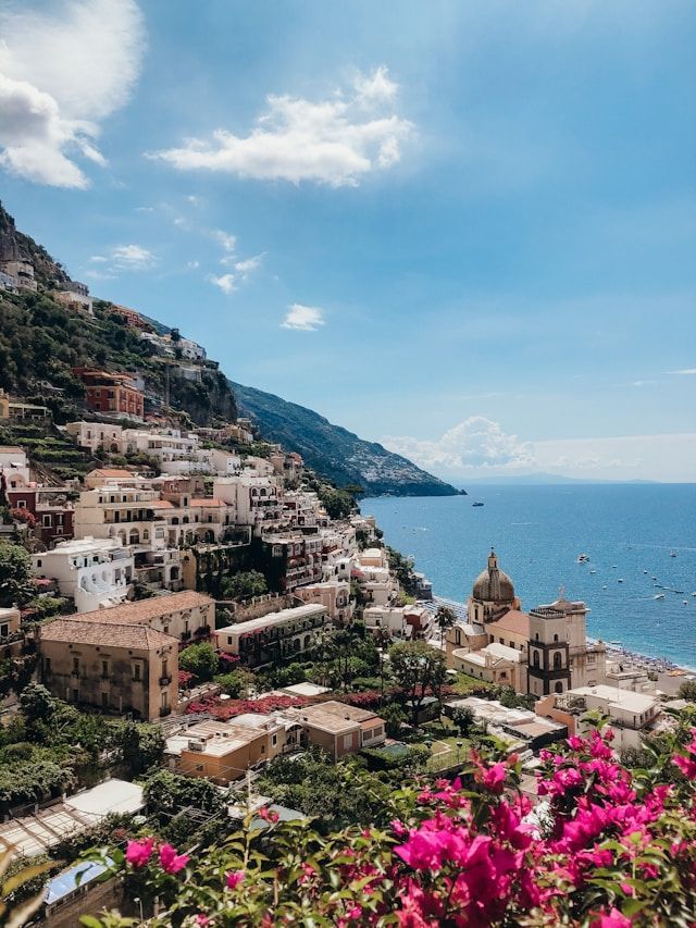 Vista elevada de un pueblo costero en un acantilado sobre el mar, con vibrantes flores rosas en primer plano.