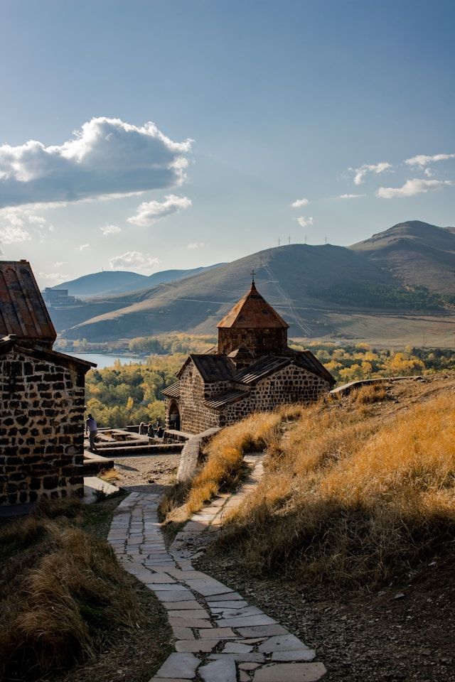 A winding stone path on a grassy hill leads to an old stone church, with mountains and a lake in the distance.