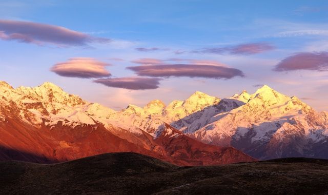 A snow-capped mountain range is illuminated by the golden light of sunrise against a blue sky with thin purple clouds.
